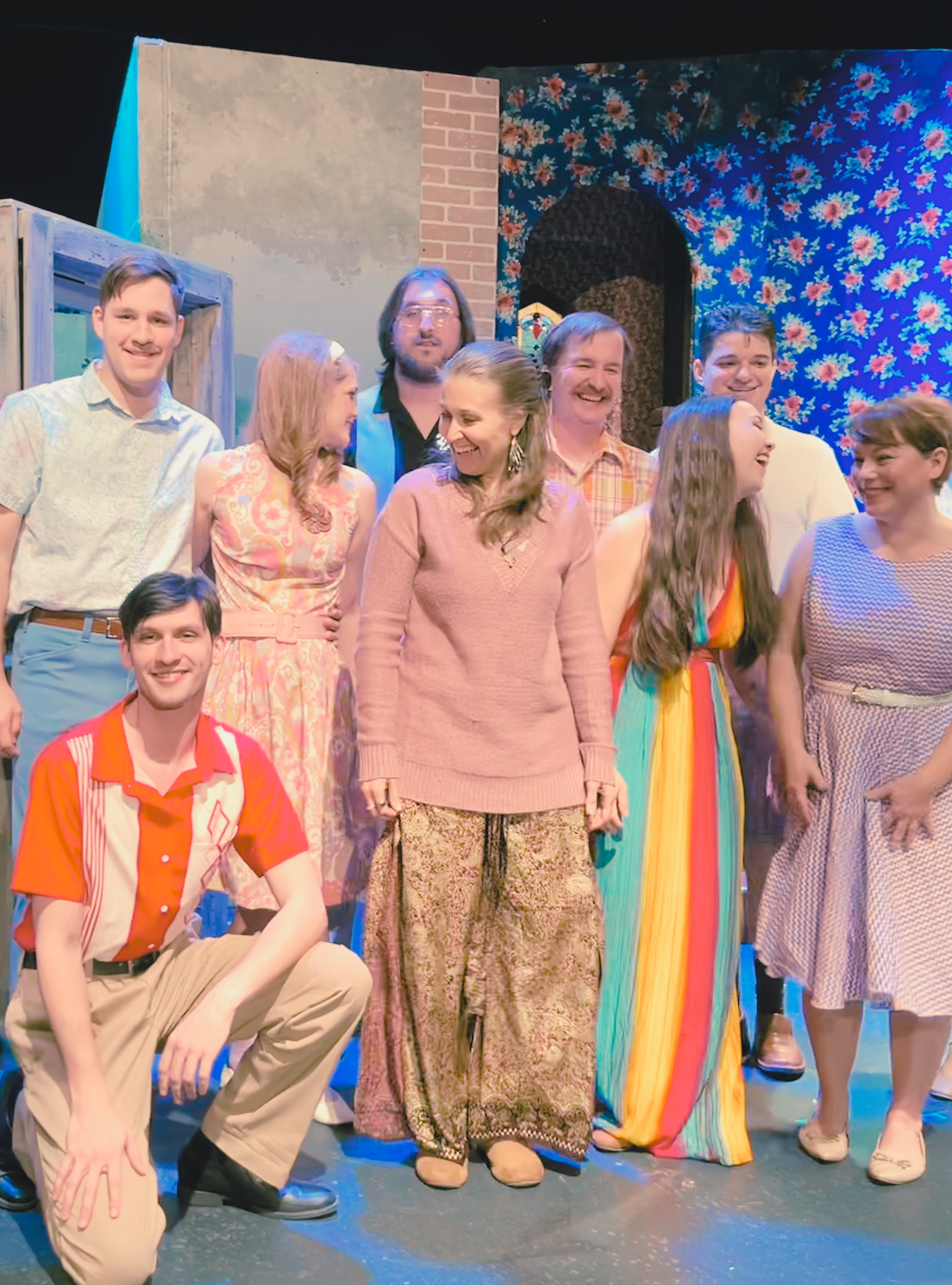 A cast of colorful characters and actors stands laughing on stage.