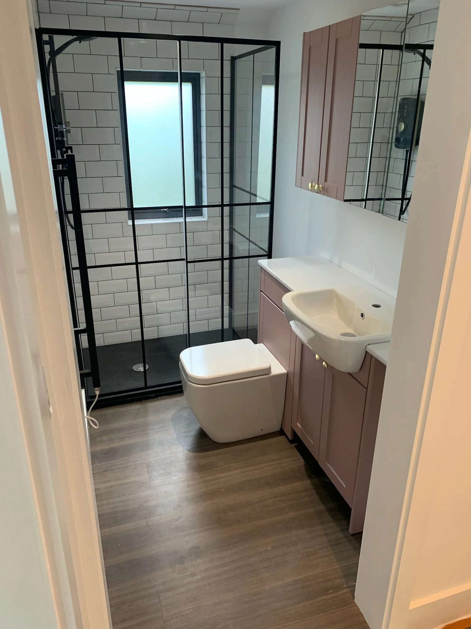A small bathroom with a shower enclosure featuring black framing, a frosted window, a white toilet, a pink vanity cabinet, a wall-mounted mirror cabinet, a white sink, and wood-look flooring.