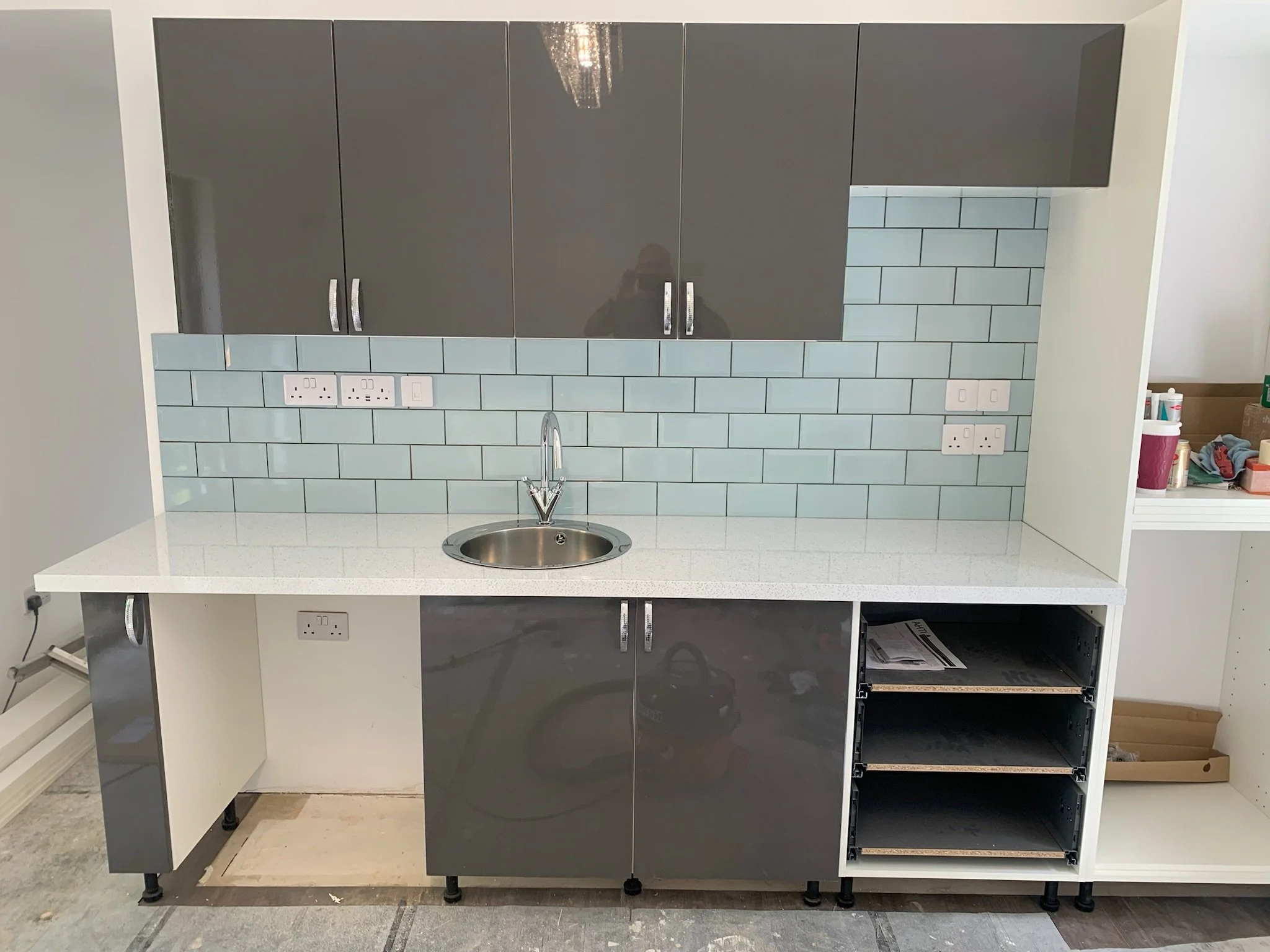 Unfinished kitchen cabinet with upper grey doors, a lower white cabinet with one door, and an open space for drawers. A blue tile backsplash and outlets are visible above the countertop. The room appears to be under construction or renovation.