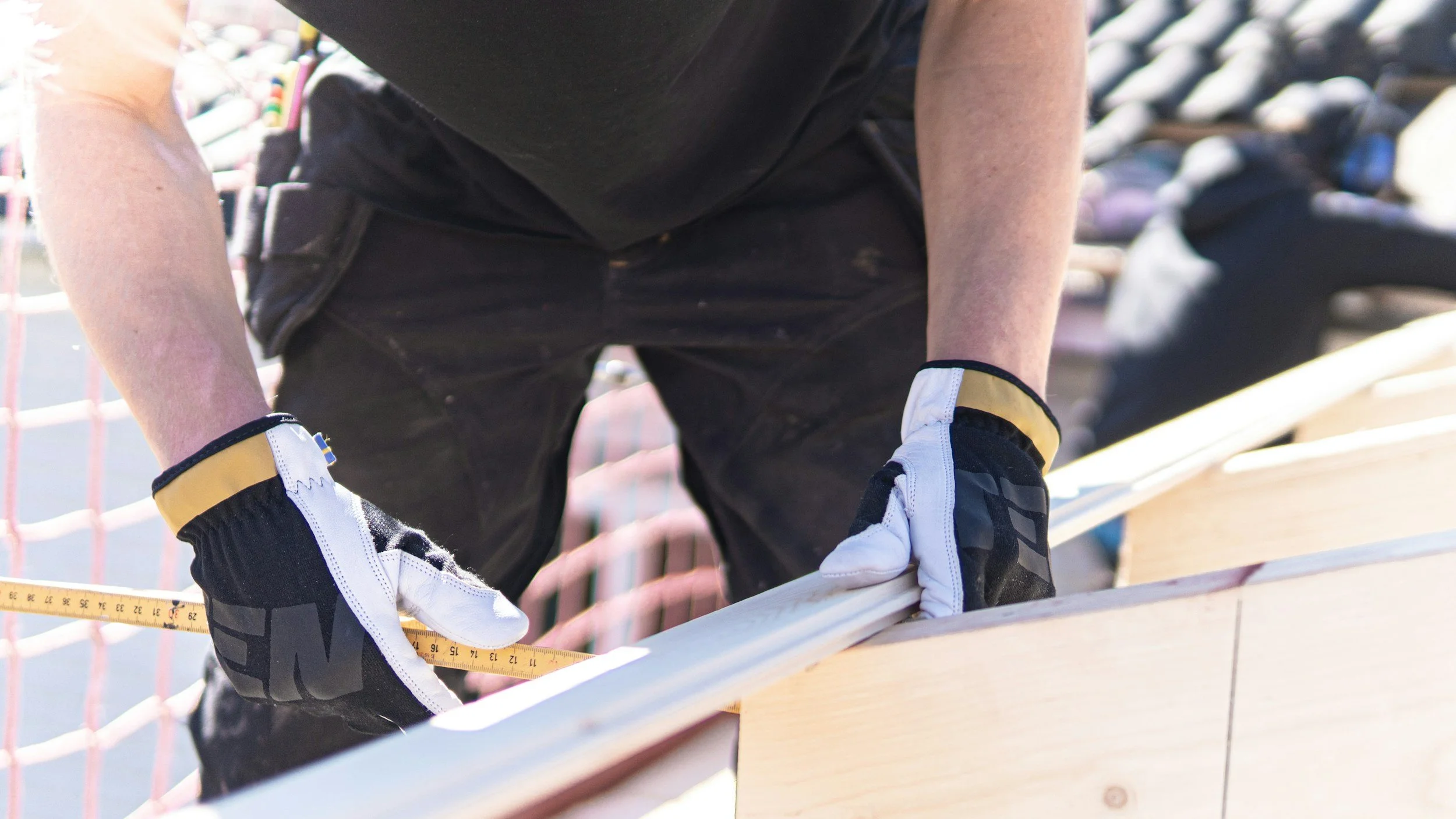 Close-up of a guernsey handyman measuring a piece of wood with a yellow measuring tape, wearing black gloves with white and yellow accents, and a black shirt.