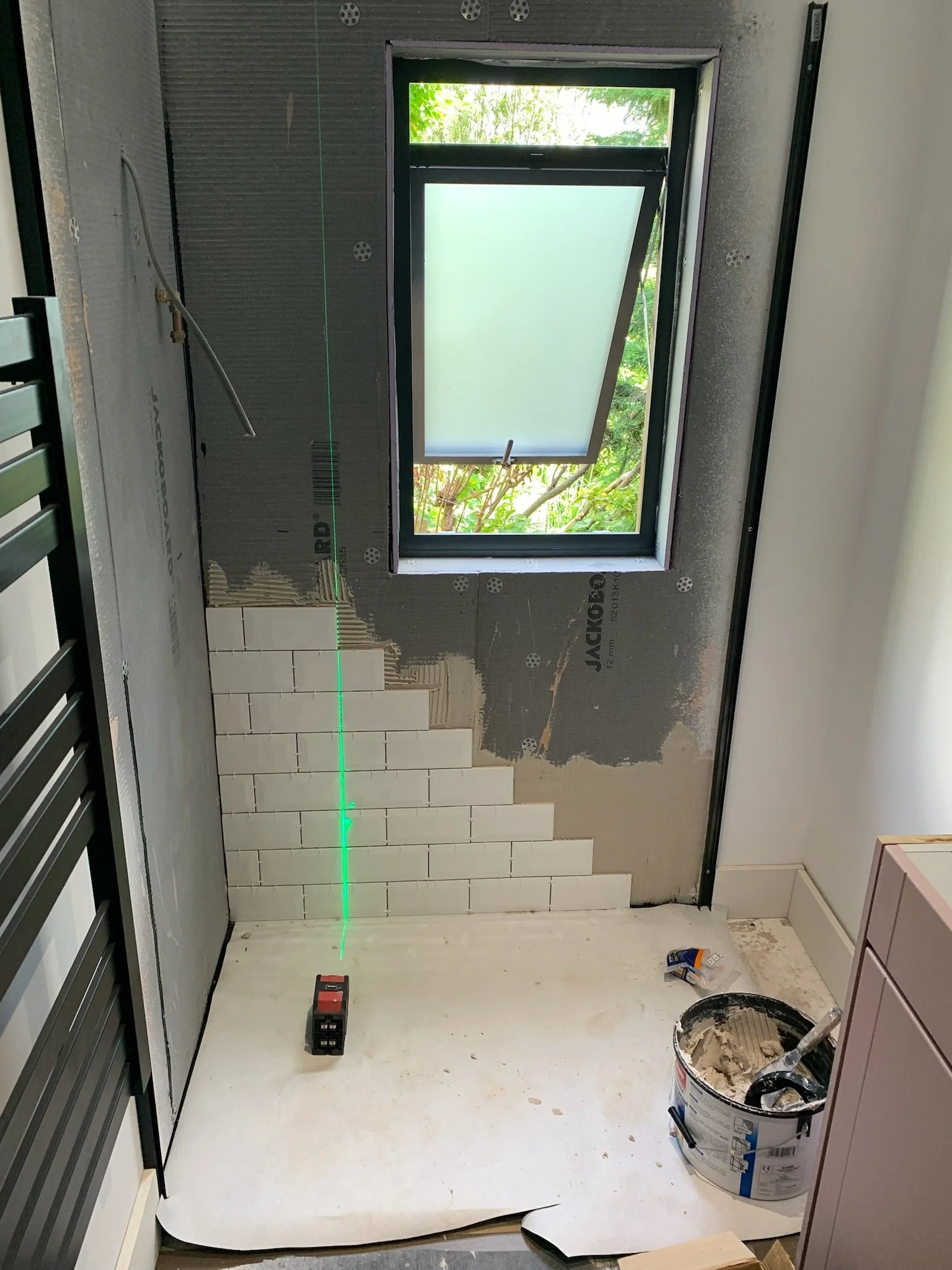 Construction interior with a window, green laser line on the floor, drywall, tools, and materials on the floor.
