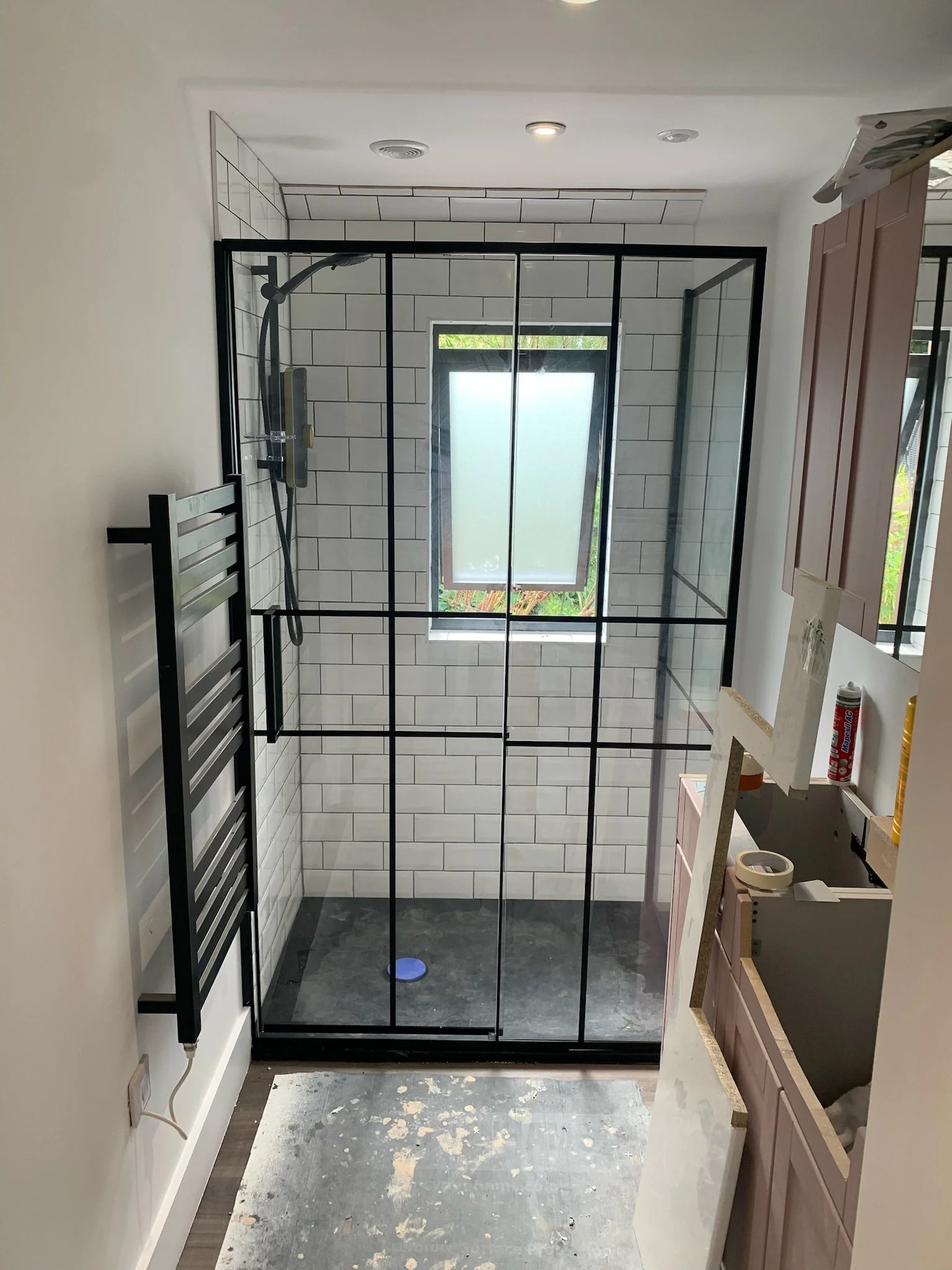 A bathroom with a glass shower enclosure, white tiled walls, and an open window. There is a black towel radiator on the left wall and construction materials and tools on the right countertop, with a filled caulk tube and a small piece of wood or dres