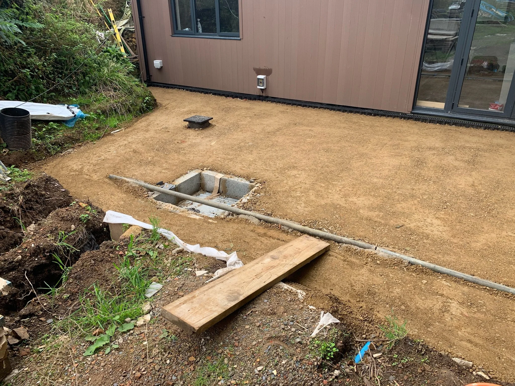 A backyard construction site in guernsey with a leveled dirt ground, a wooden plank, an outlet on the house wall, and a nozzle on the ground. There's a small square drain or vent and some construction materials and tools on the side.