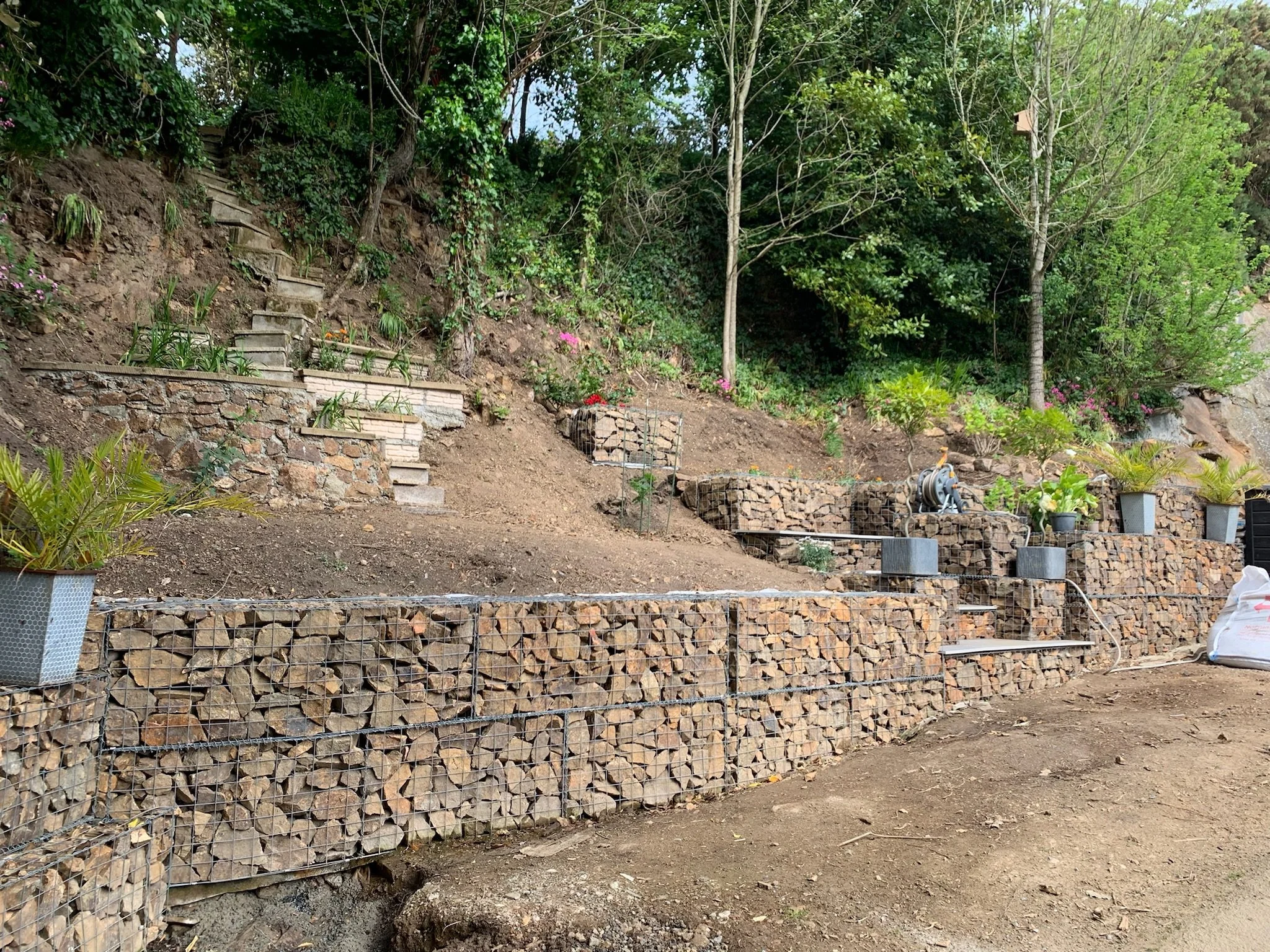 A hillside garden under construction with stone steps, retaining walls made of landscape rock, flower pots, and trees with green foliage.