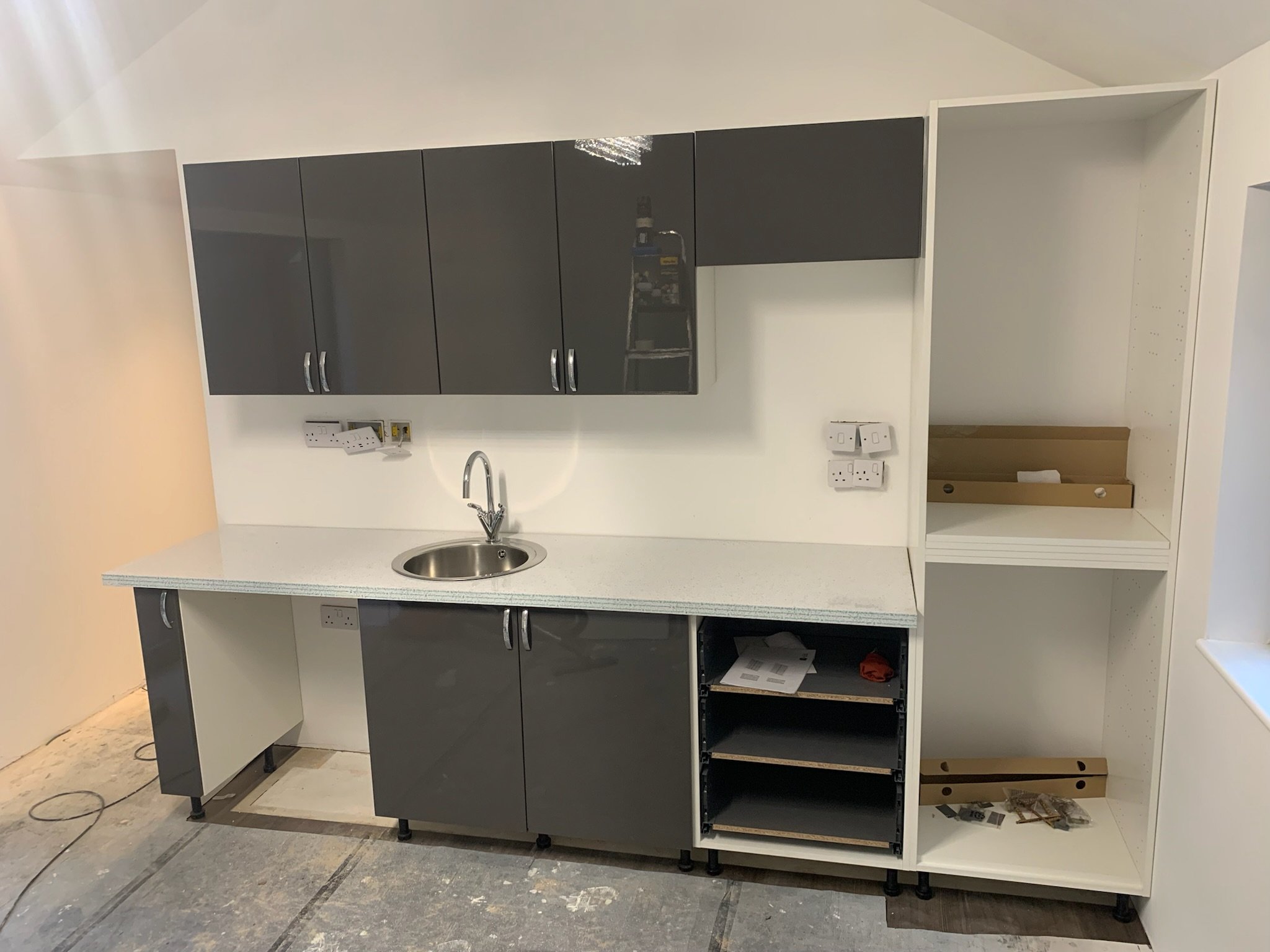 Kitchen cabinets installation in progress, with upper black cabinets and an open white cabinet, a small round sink, and electrical outlets on the wall.