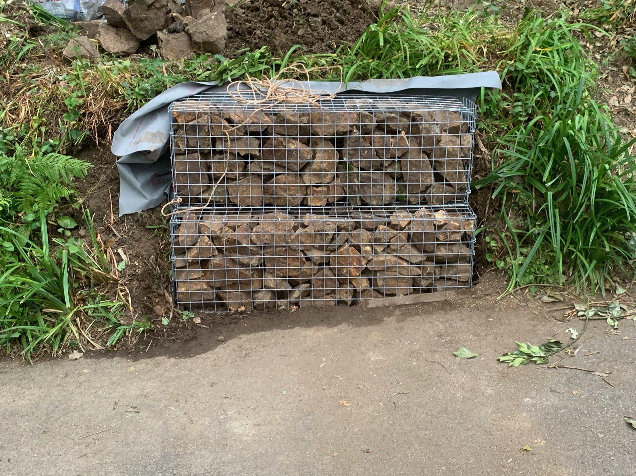 A wire mesh container filled with large rocks, placed on the ground next to a patch of green grass and foliage, with a pile of dirt and rocks behind it.