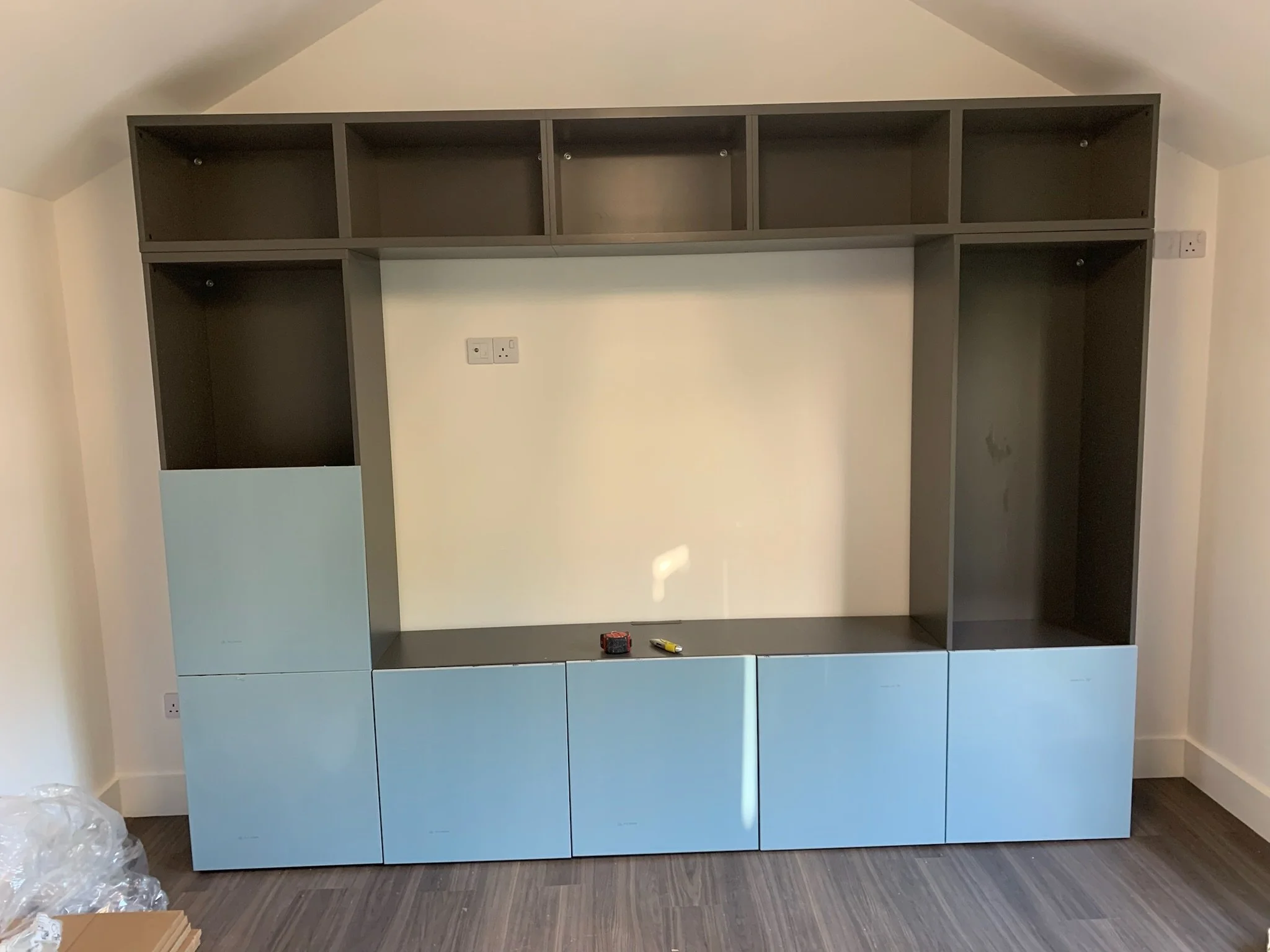Unassembled gray and light blue shelving unit mounted on a white wall in a room with wooden flooring, with tools placed on the bottom shelf.