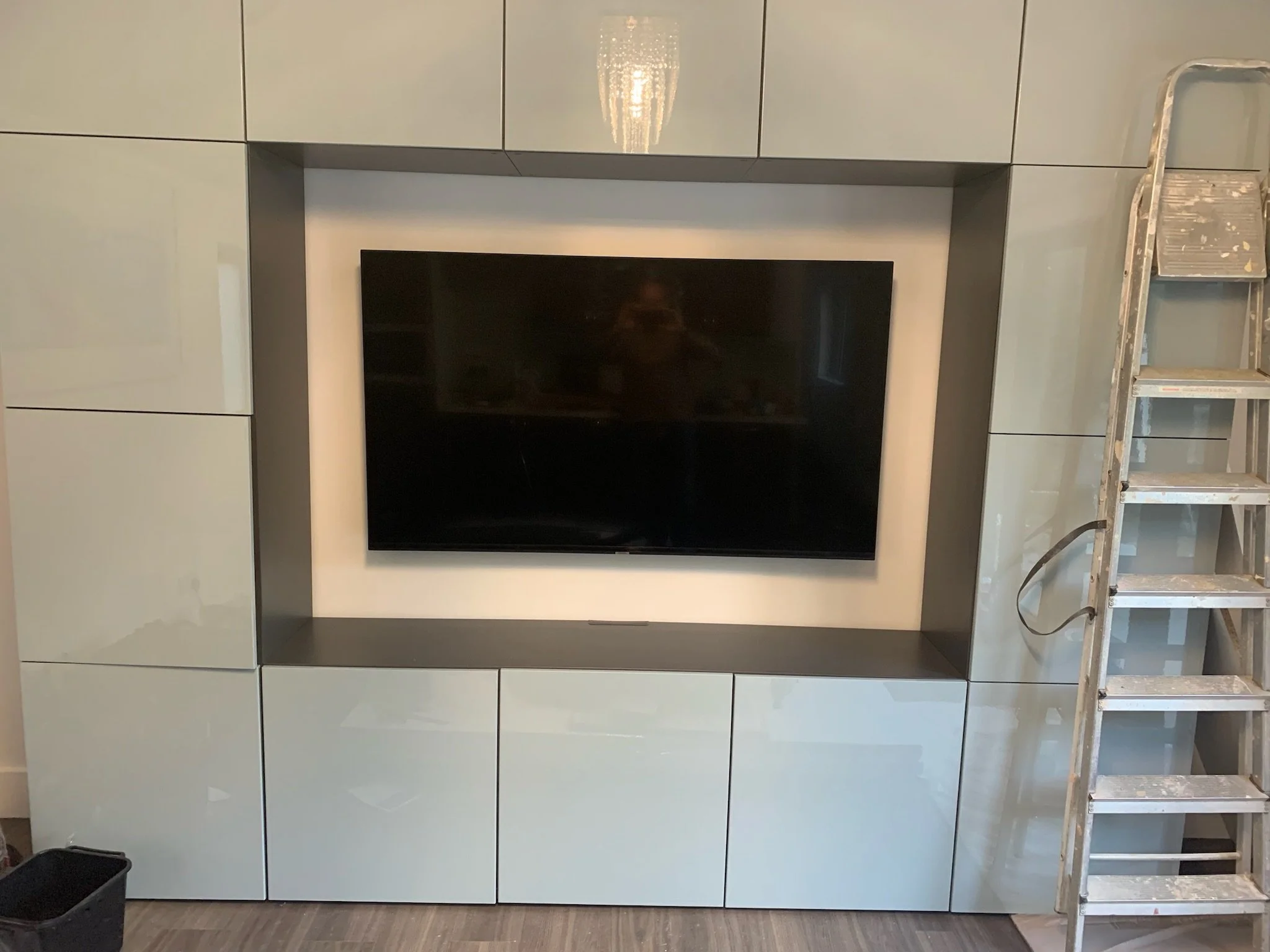 A partially assembled built-in wall unit with closed cabinets, a TV niche, and a ladder leaning against the right side, in an indoor space.