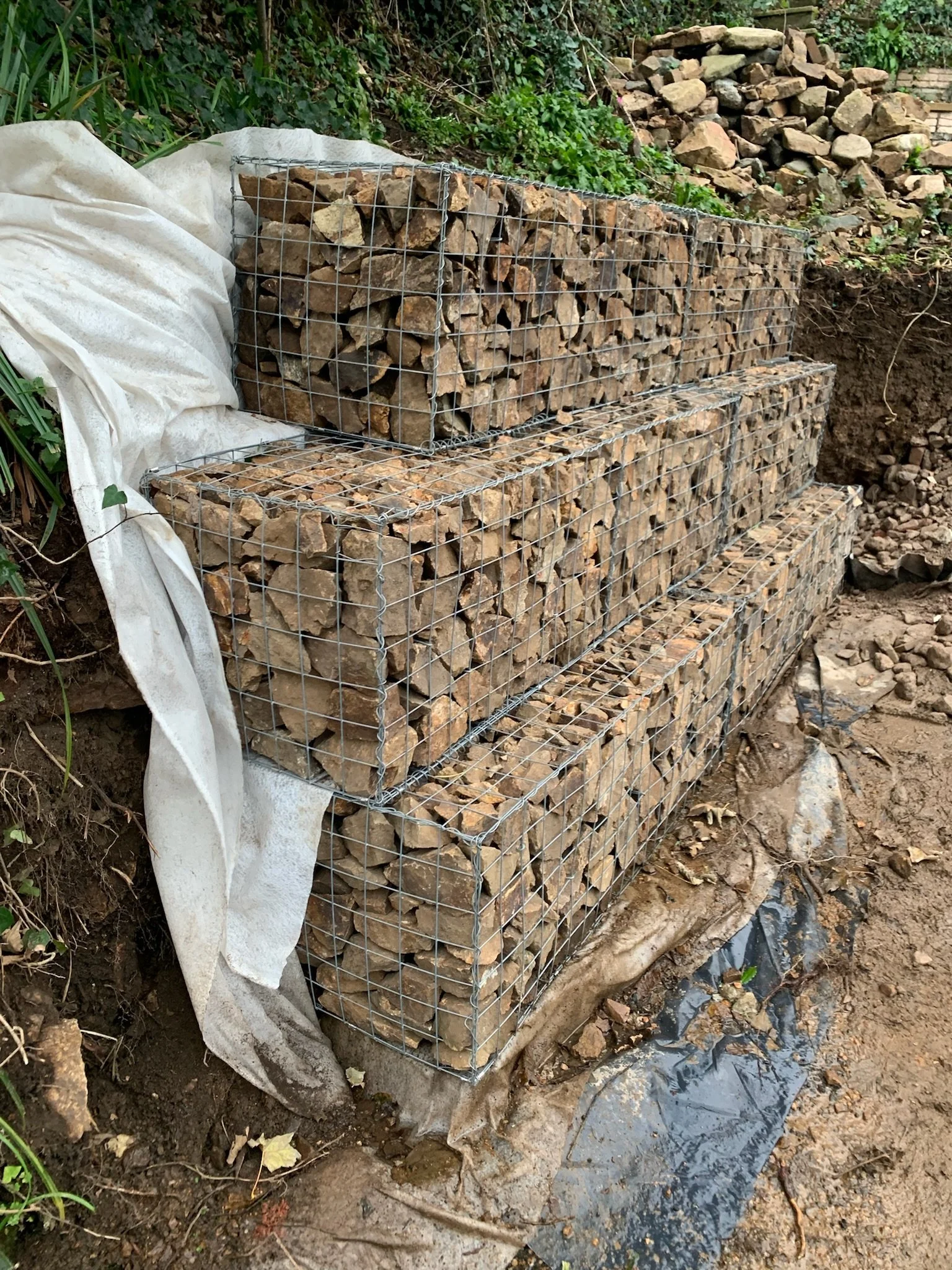 Stacked rocks enclosed in wire mesh for retaining wall, on a slope outdoors with some greenery and soil nearby.