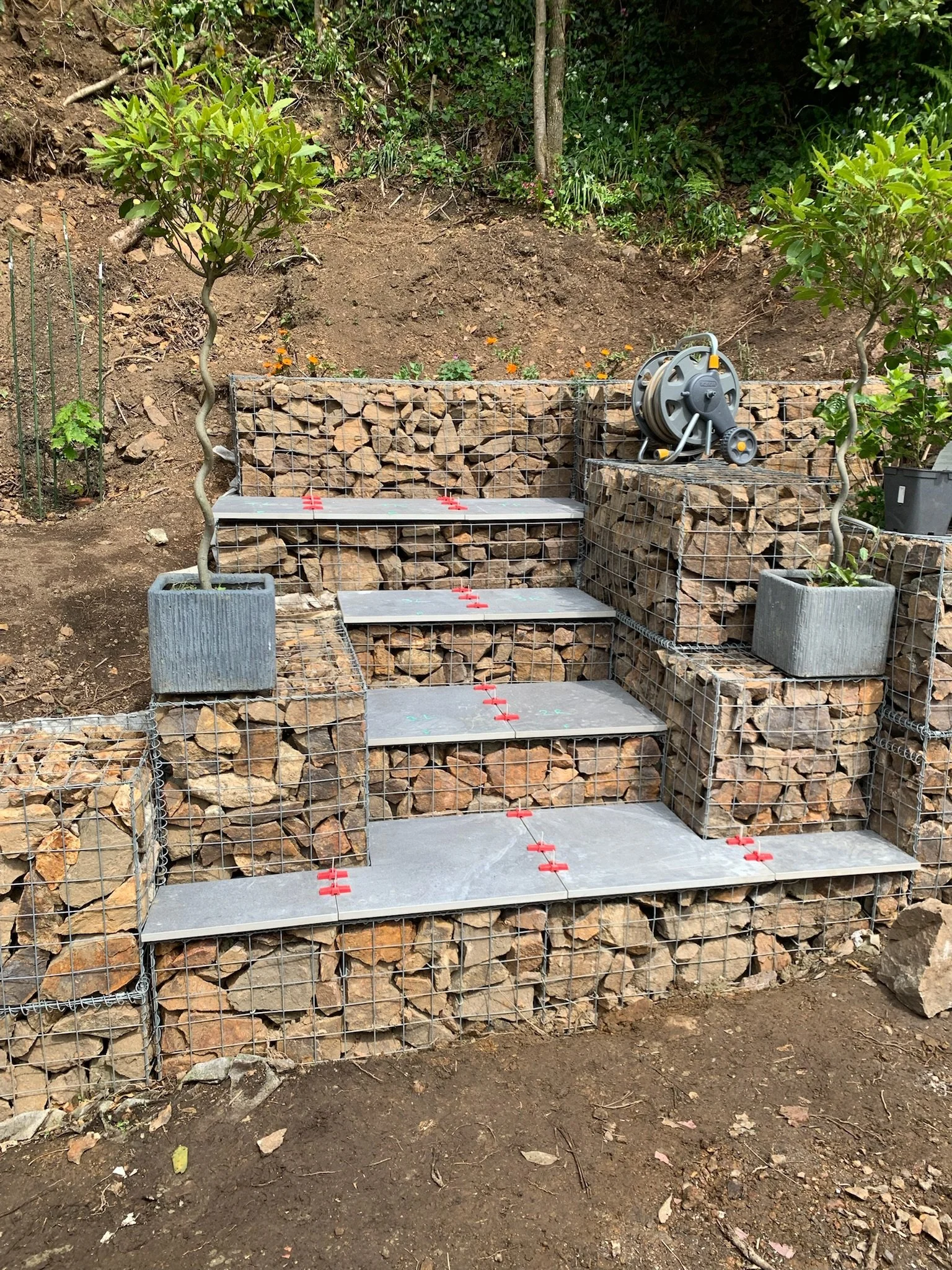A stepped garden feature with metal mesh sides filled with rocks, with a hose reel on top and two potted plants flanking it, set in an outdoor garden area with soil and plants in the background.