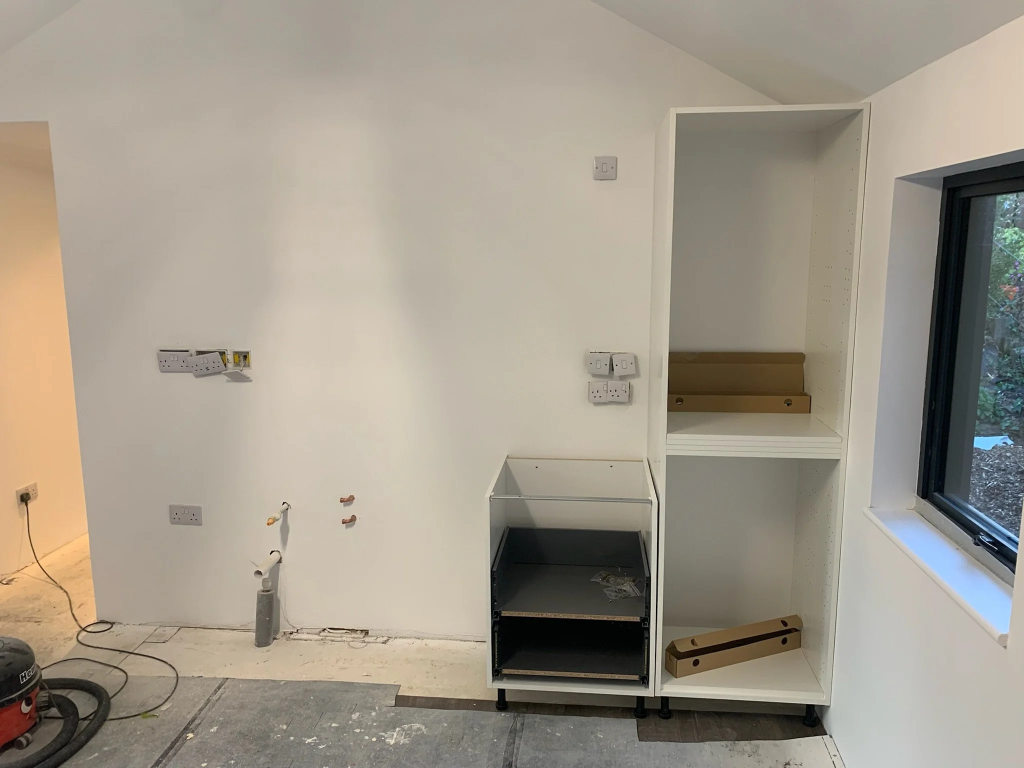 White wall in a room under construction with wall sockets, pipes, and an unfinished built-in shelf or cabinet, next to a window showing outdoor greenery.