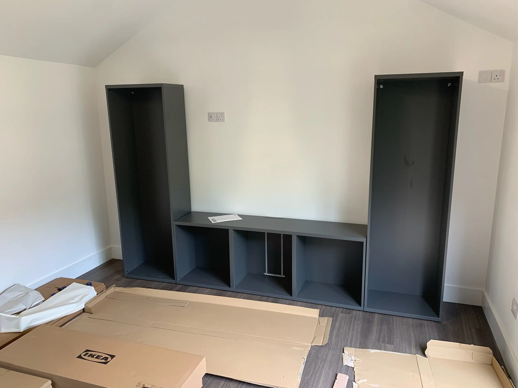 A partially assembled black shelving unit in a room with white walls and dark flooring, with IKEA boxes and packing materials on the floor in front of it.