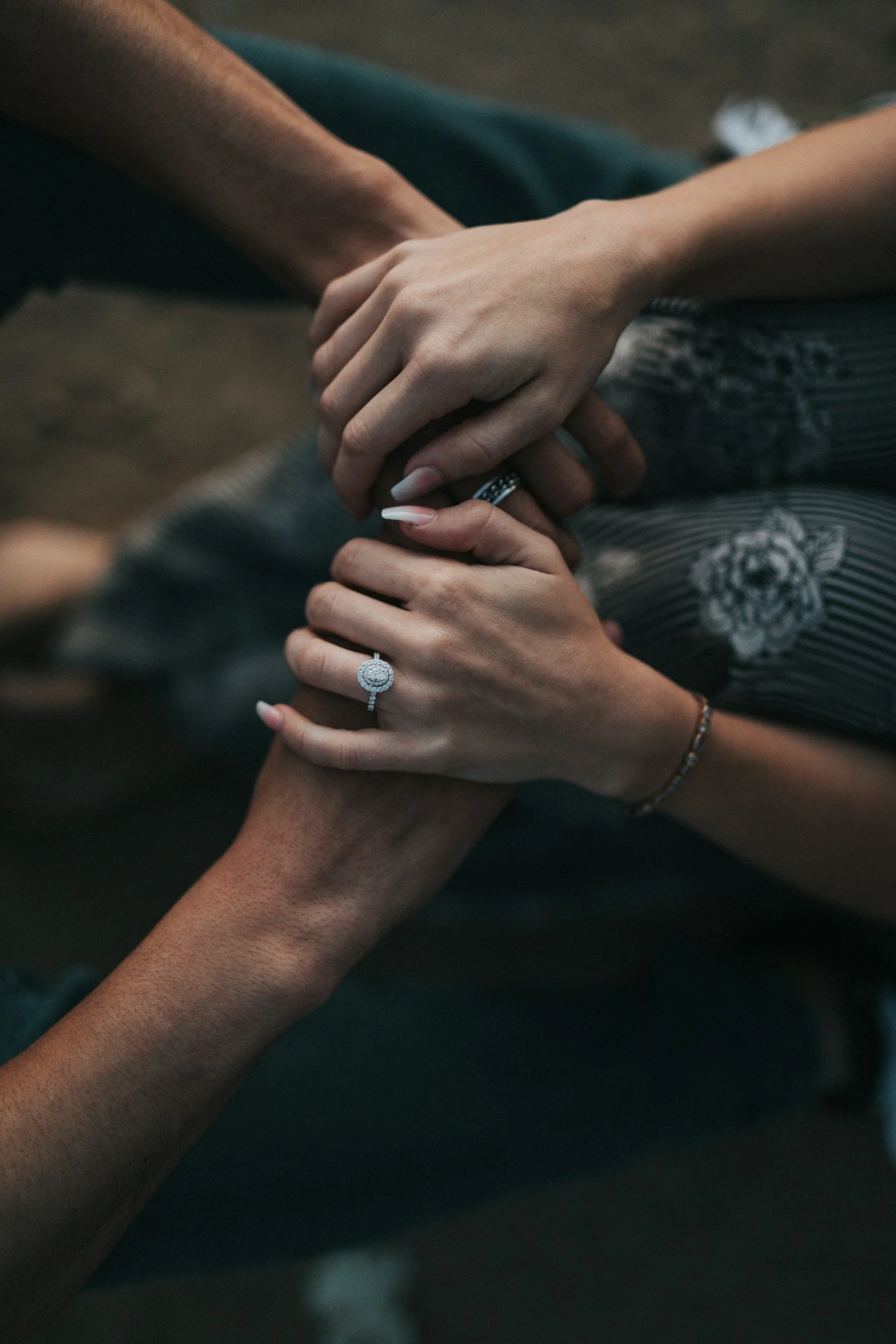 A close-up of four hands gently holding each other, displaying rings on their fingers, with dark clothing and a dark background.