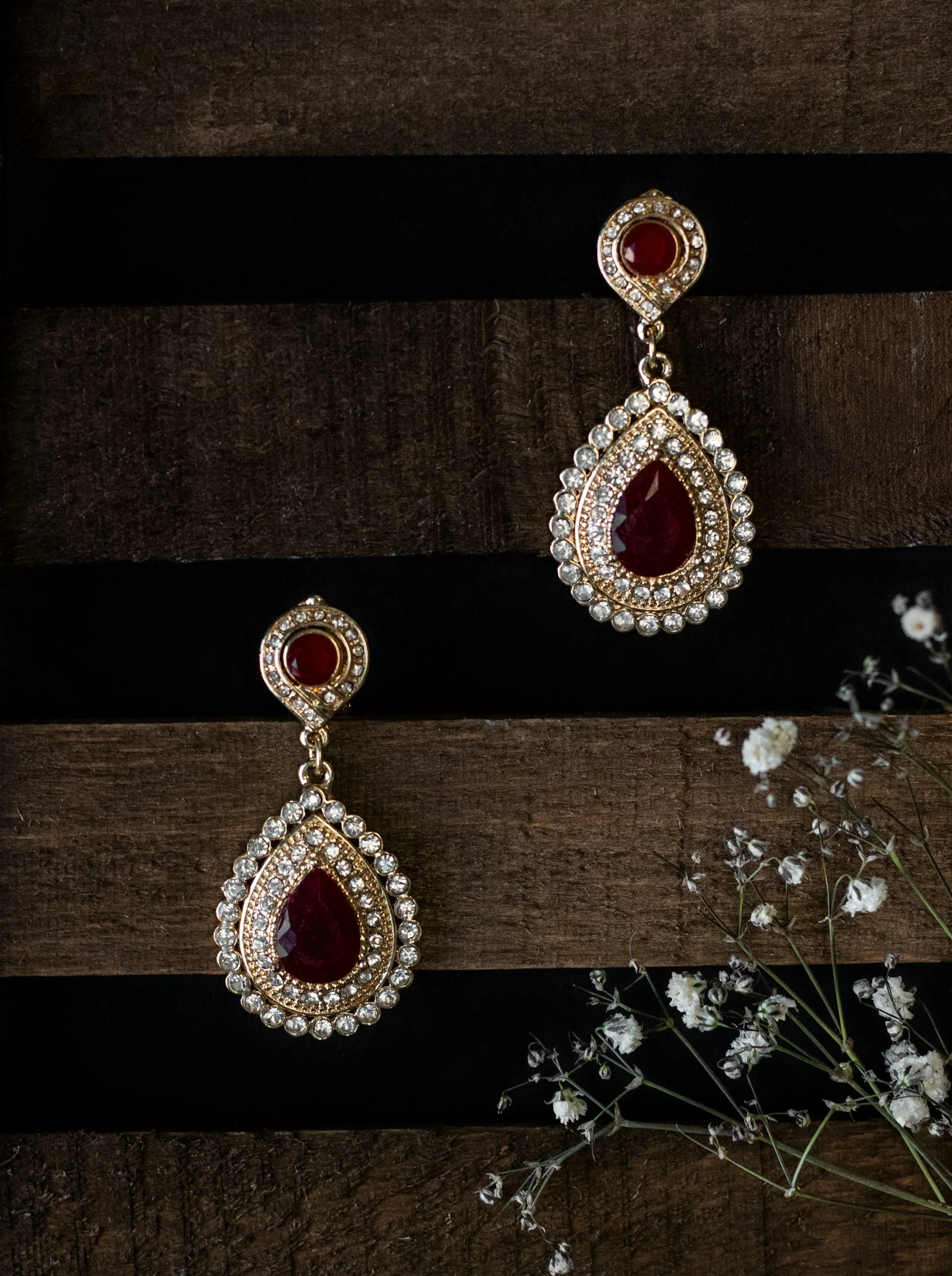 A pair of gold earrings with large red gemstones and surrounded by clear rhinestones, displayed against a dark wooden background with white small flowers on the side.