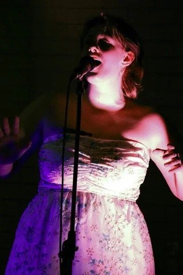A woman singing into a microphone on stage, illuminated by pink and purple lights, wearing a strapless floral dress.