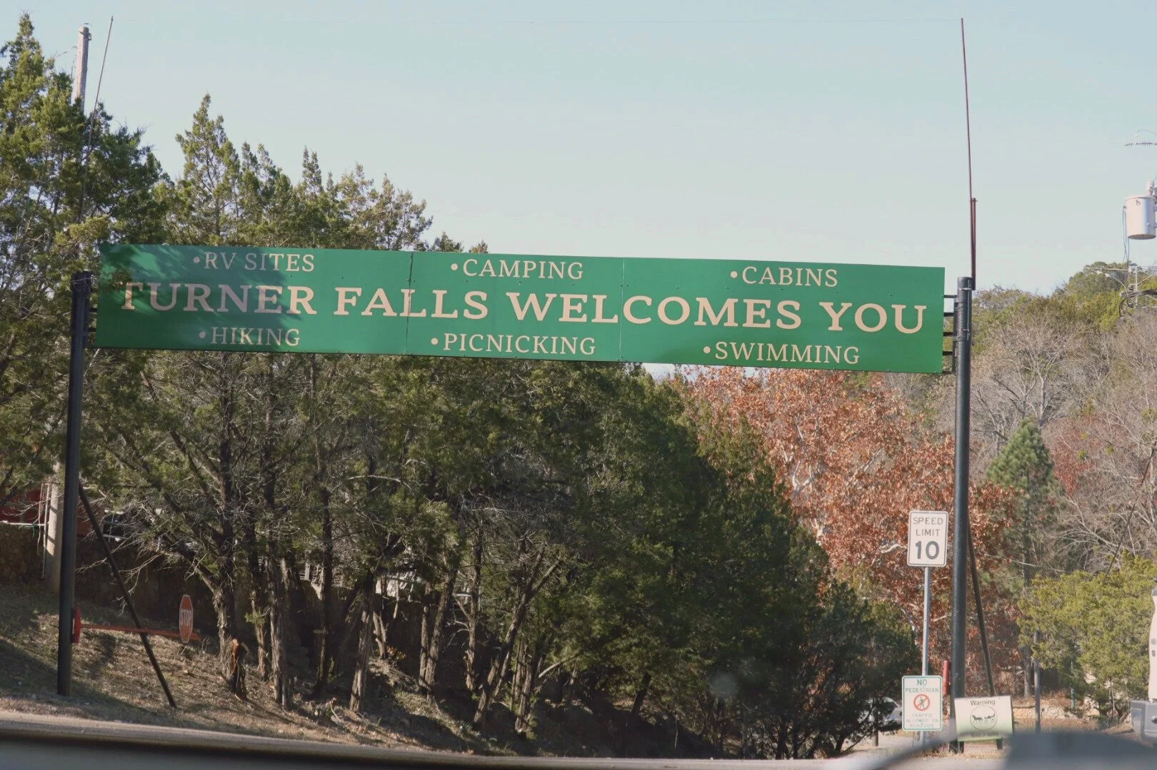 Green roadside sign that reads 'Turner Falls Welcomes You' with additional text listing activities and amenities such as RV sites, camping, cabins, hiking, picnicking, and swimming. Trees and a speed limit sign are visible in the background.