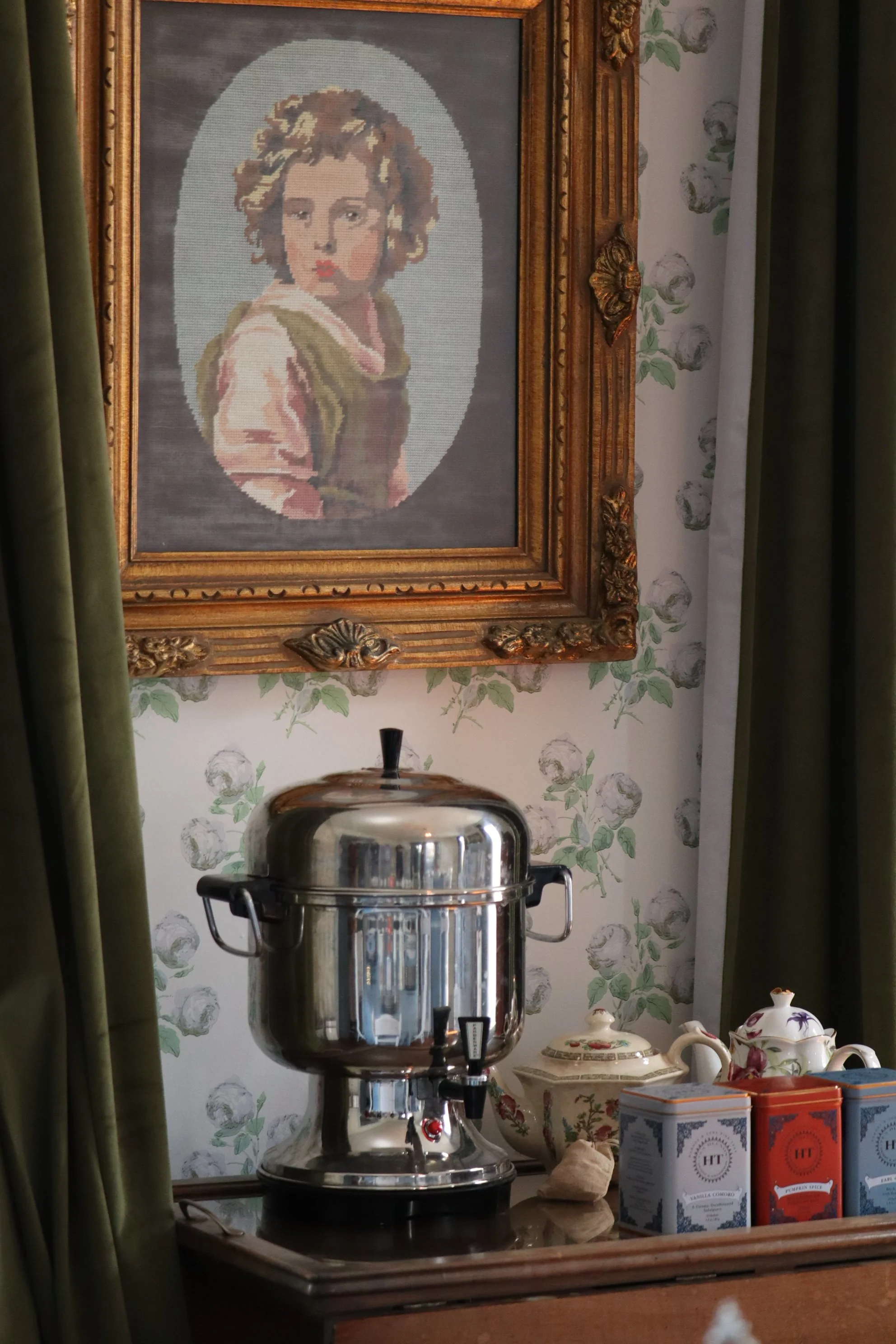 A framed cross-stitch portrait of a woman with curly hair, placed on a wall with floral wallpaper. In the foreground, there is a stainless steel coffee urn, and to the right are decorative teapots and boxes of tea on a wooden surface. Green curtains are partially visible on the left side.