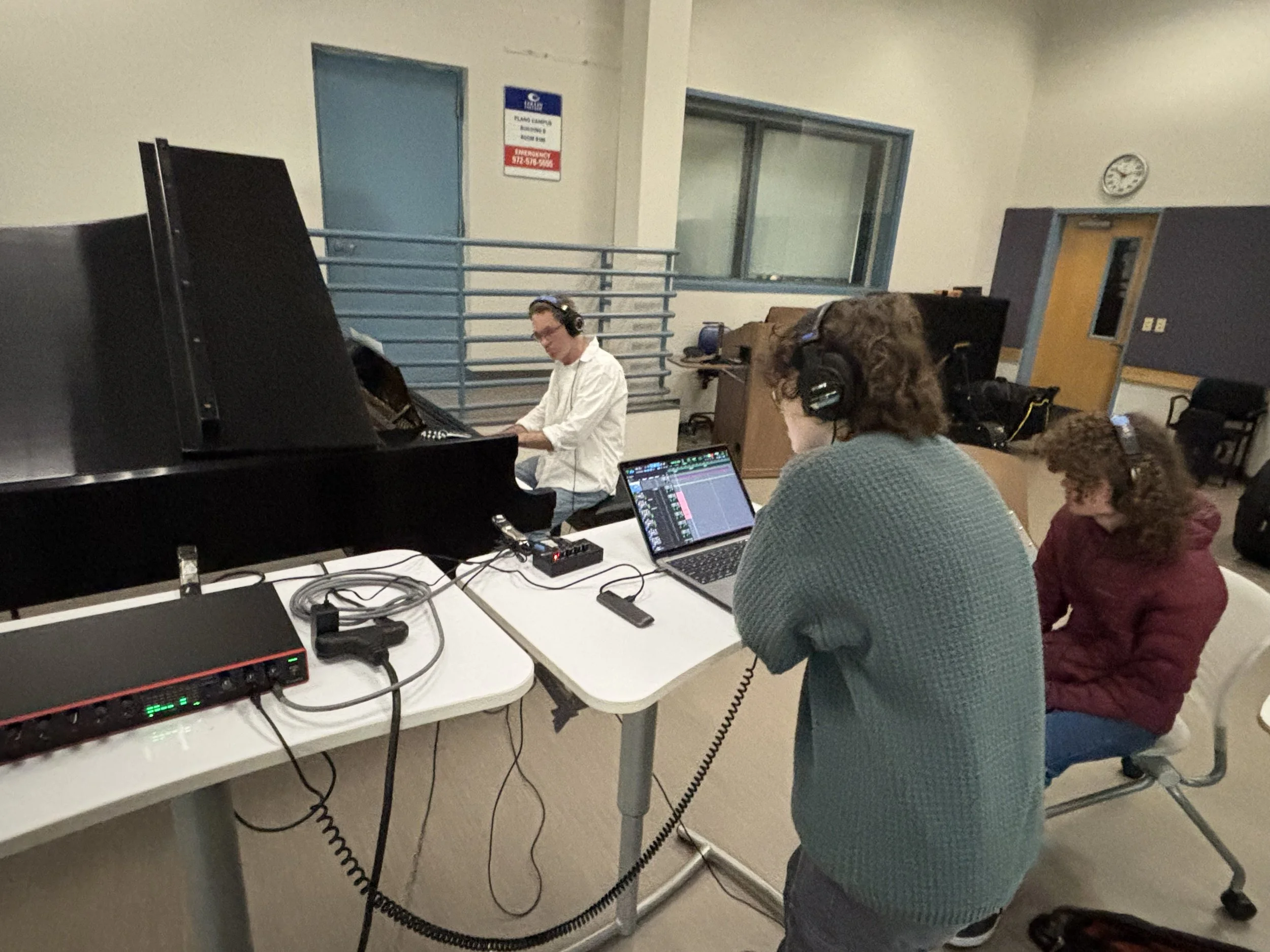 A group of people are in a room with computers and audio equipment. One person is playing a piano while others observe and work on their laptops. The room has a clock on the wall and a staircase in the background.