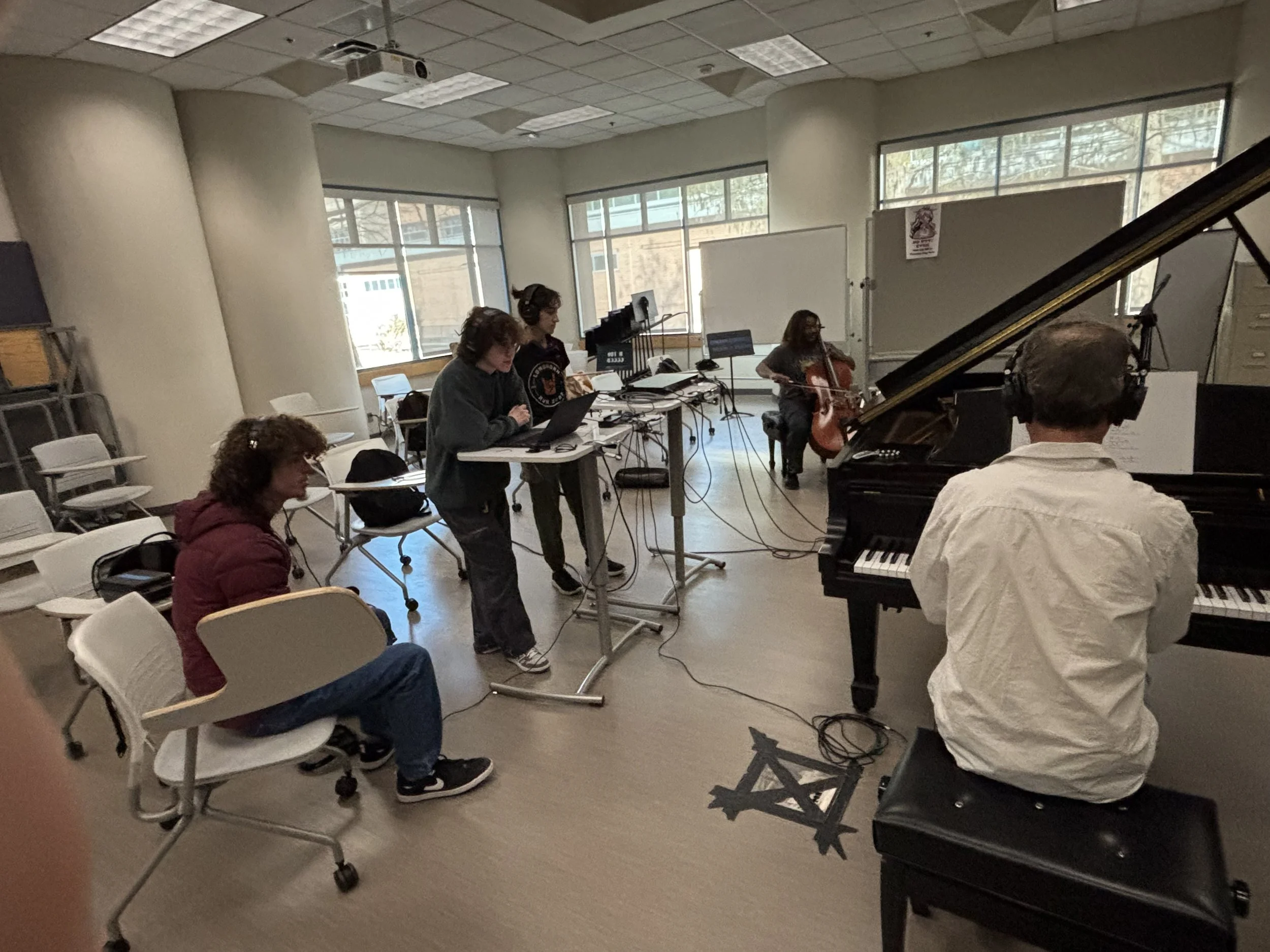 A music studio room with people playing instruments and working on computers, with large windows letting in natural light. Madeline is sat in front of a laptop with an audio engineering program open.