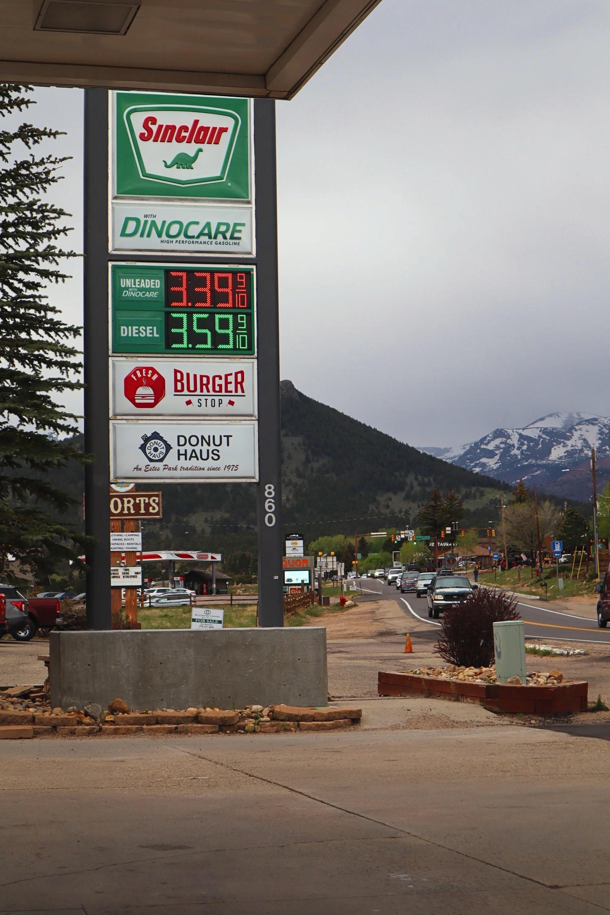 A gas station sign displaying fuel prices, with scenic mountains and trees in the background on a cloudy day.