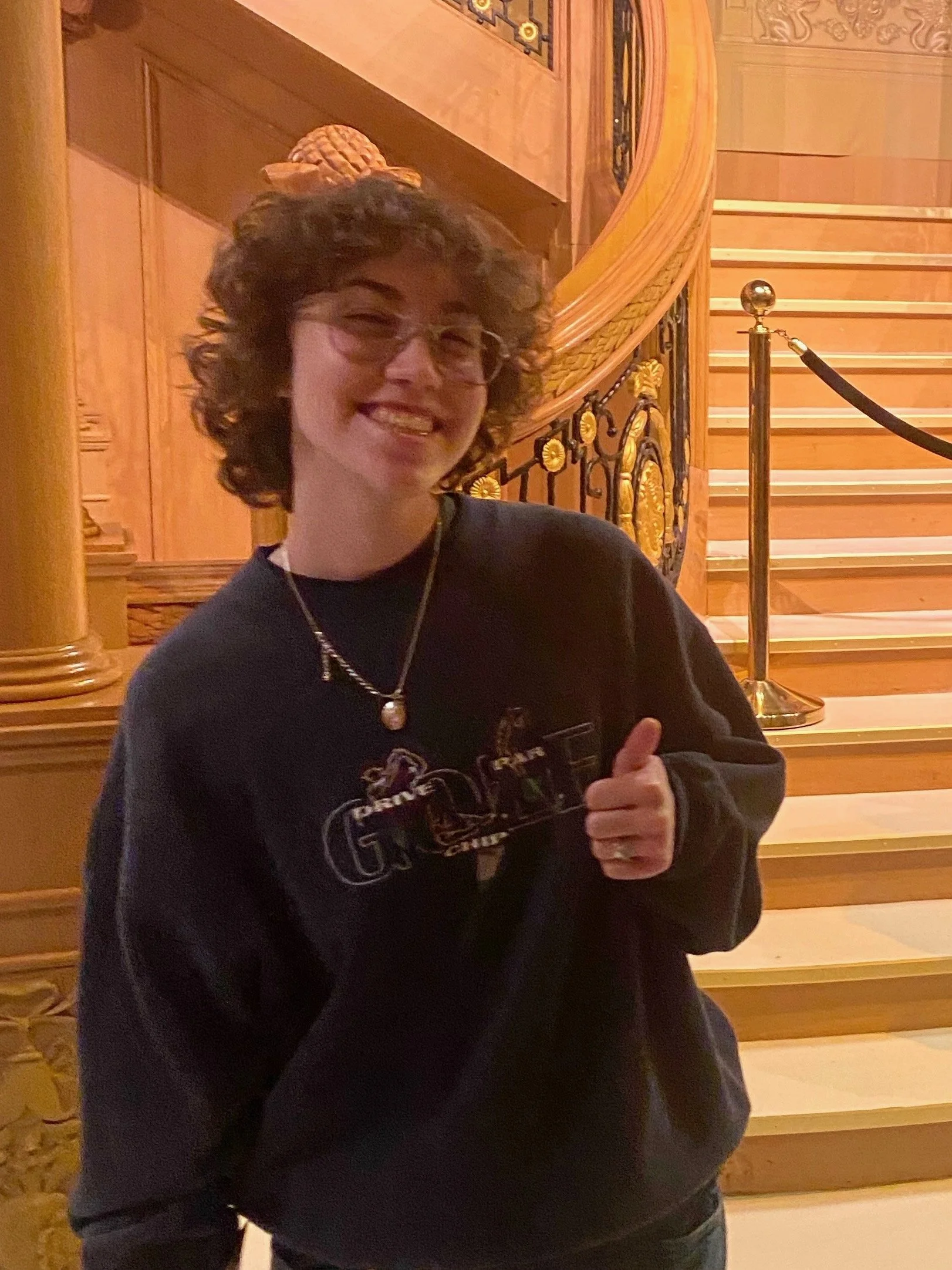 A photo of Madeline. She has curly hair and glasses, is smiling, giving a thumbs-up, in front of a staircase with gold and black railing.