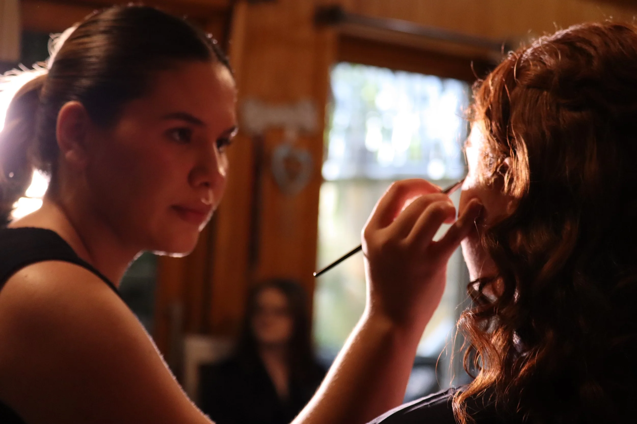 A young woman with dark hair styled in a ponytail applies makeup to another woman with curly red hair, in a warmly lit indoor setting.