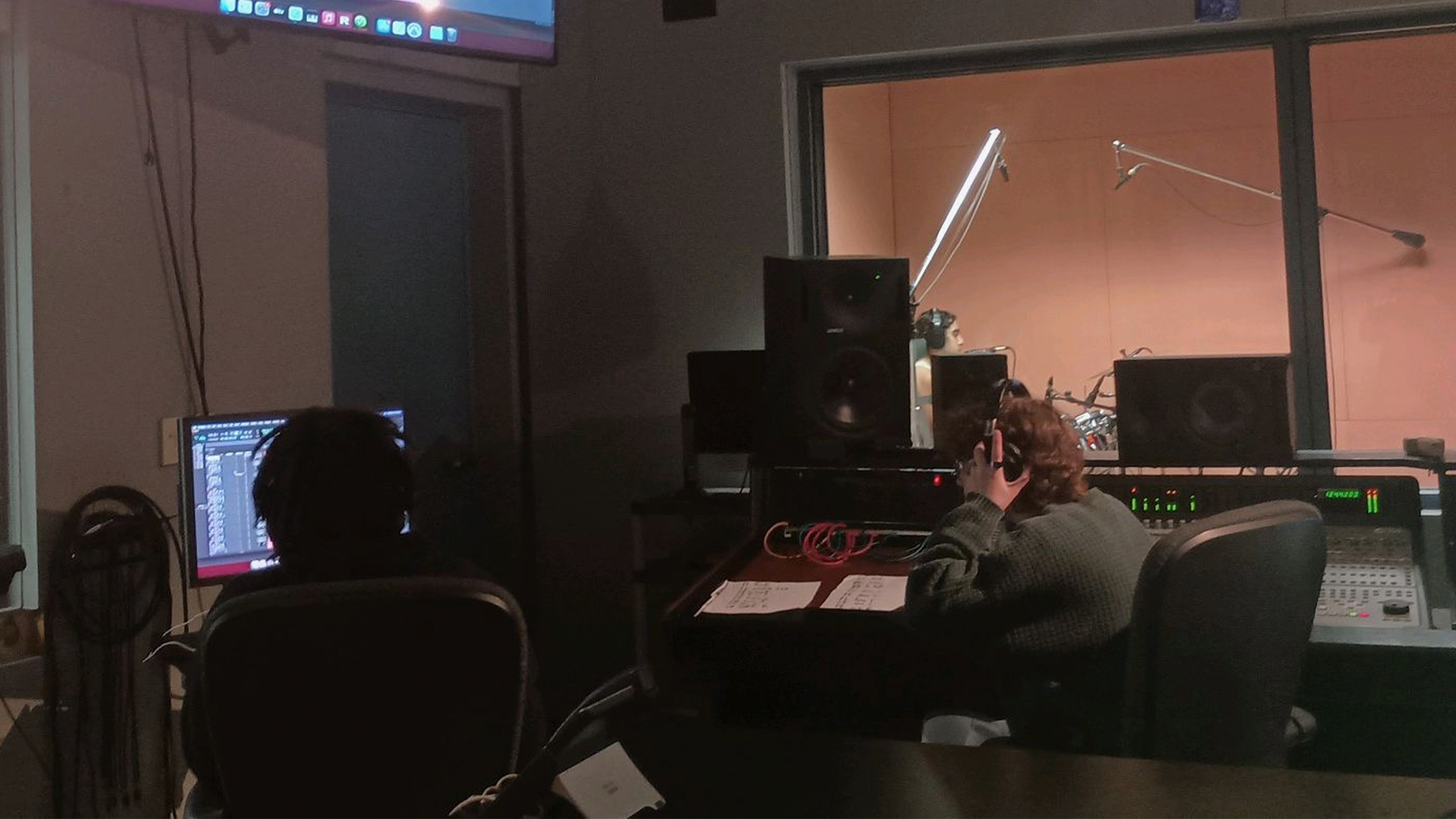 A recording studio with Madeline and a coworker working at mixing consoles. Madeline is wearing headphones, while another person is looking at a computer screen. There is a window showing a soundproof room with a person recording vocals.