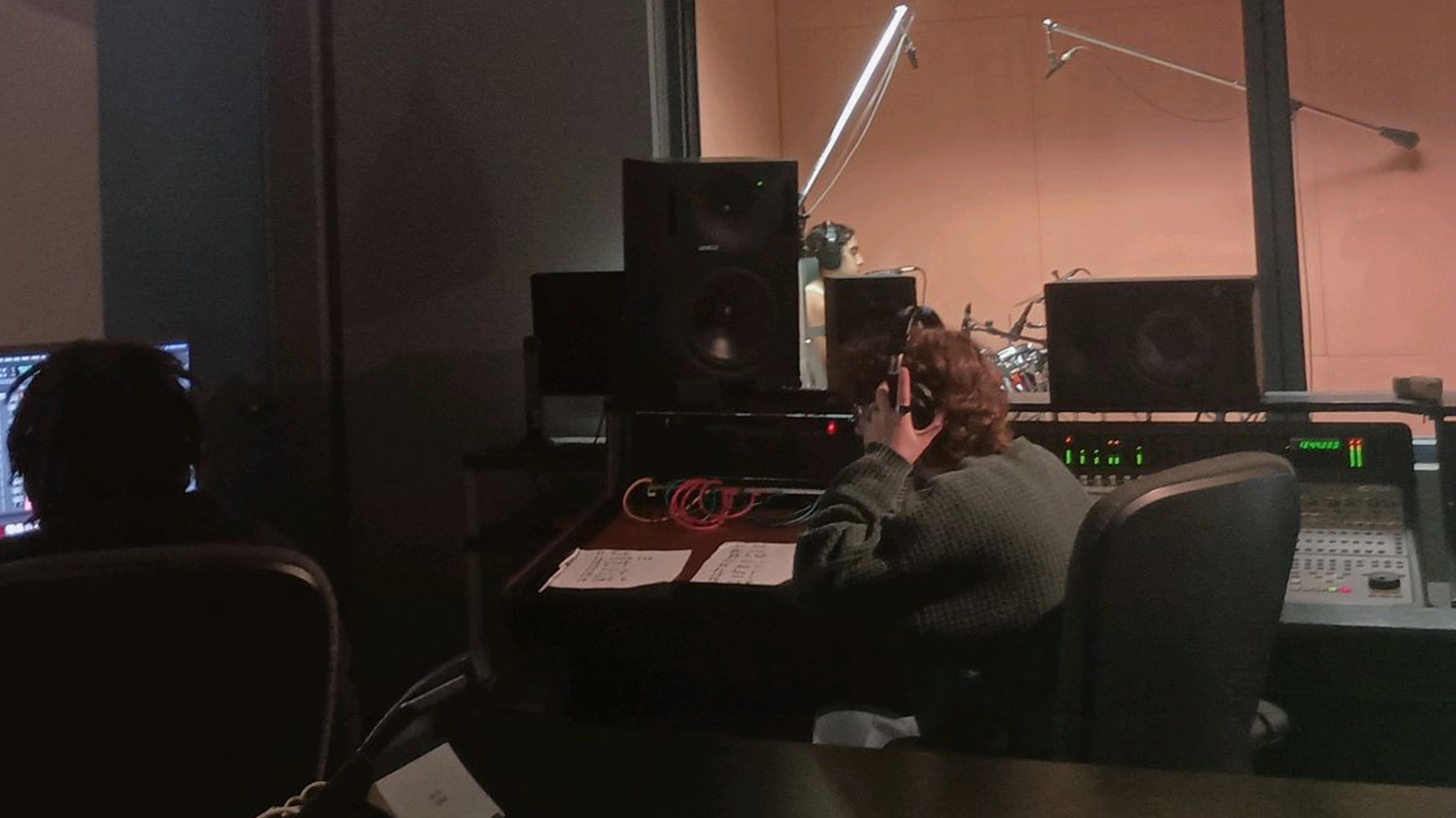 People working in a recording studio with audio equipment, monitors, and a person wearing headphones.