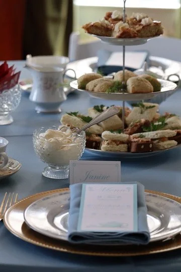 Elegant afternoon tea table with sandwiches, scones, and desserts, featuring a tiered tray and fine china. A menu sits atop the set plates.