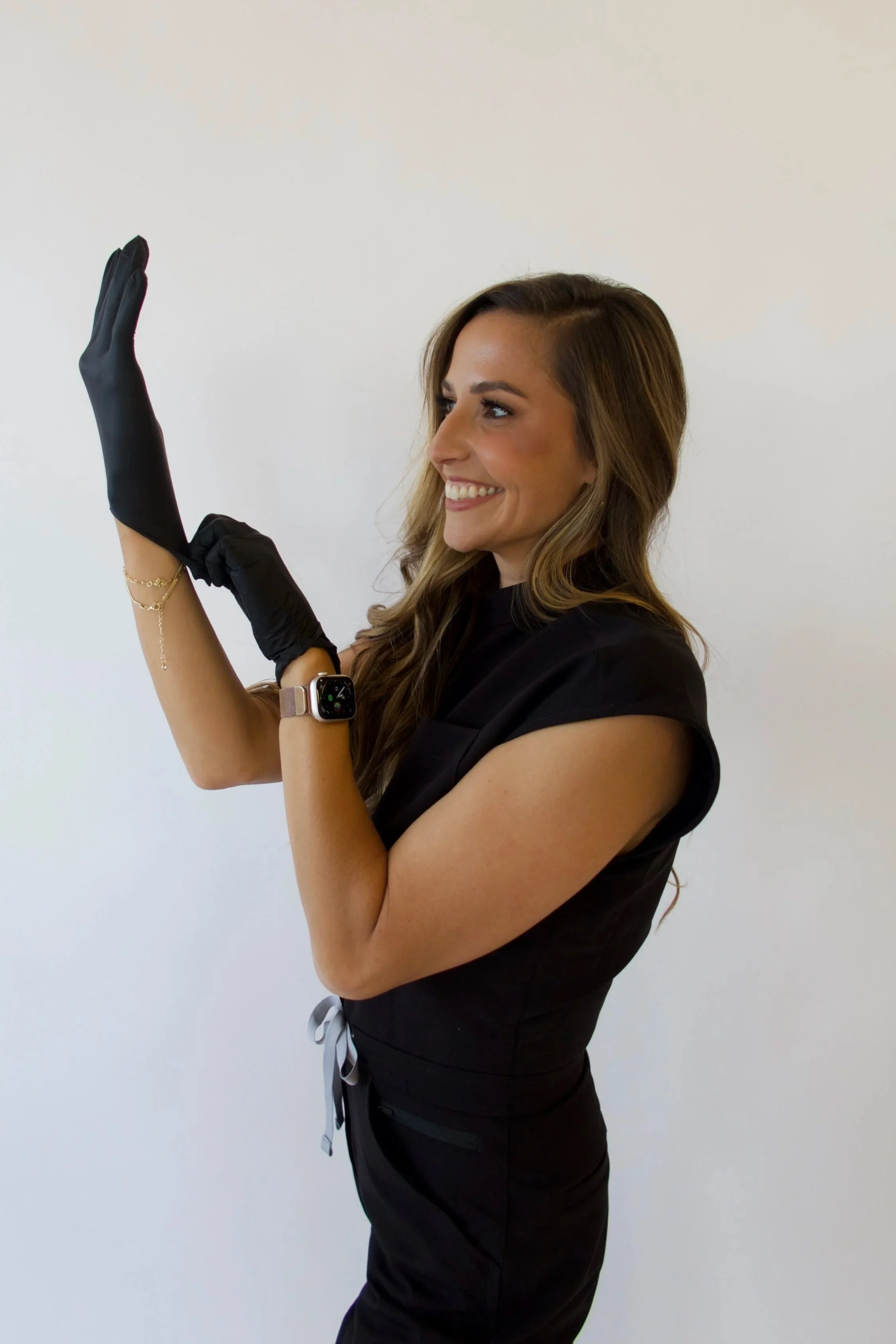 Smiling woman wearing black gloves, athletic wear, and a smartwatch warming up with arm stretches in a white room.