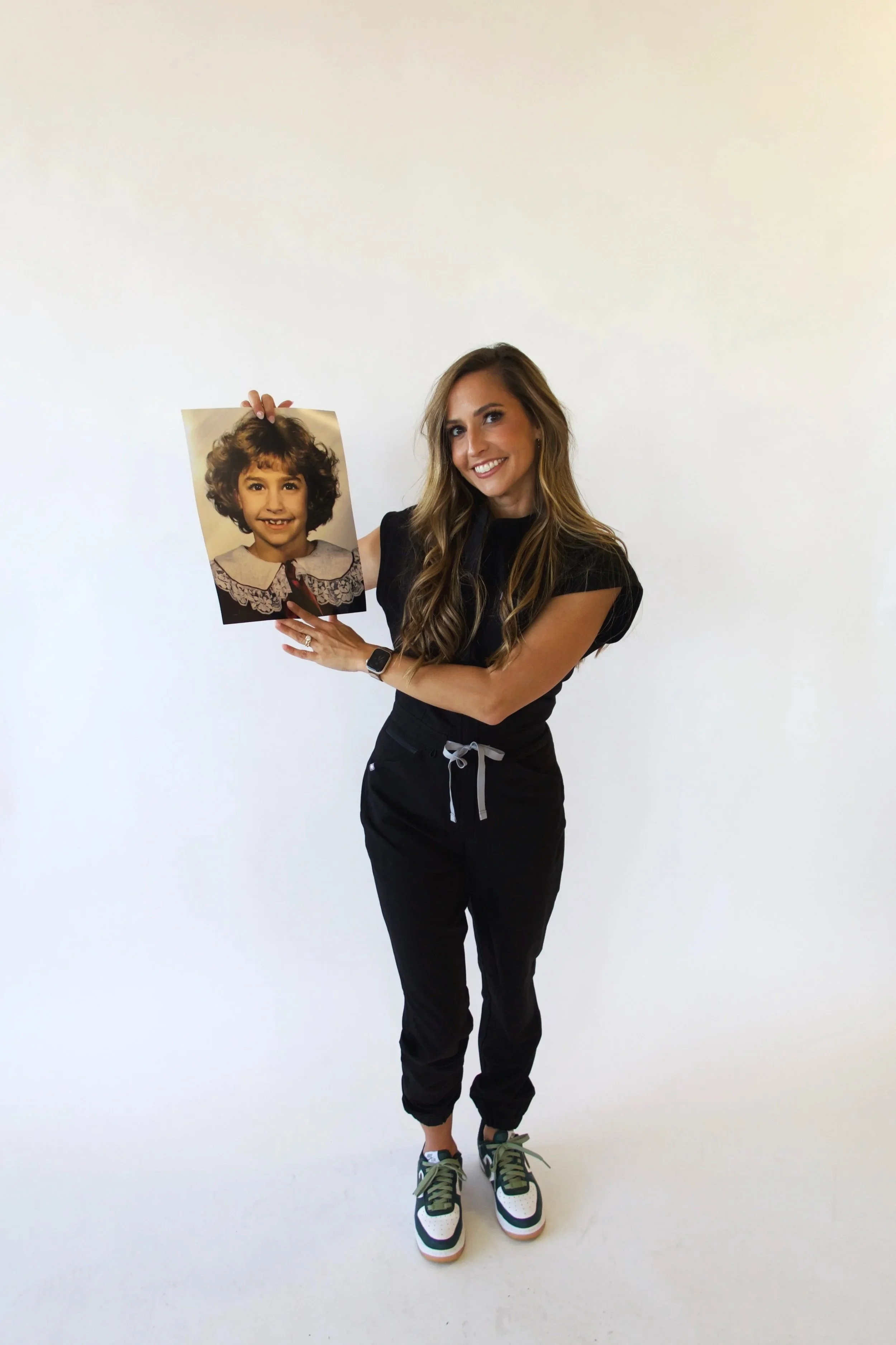 A woman with long wavy hair, wearing a black top, black pants, and sneakers, is standing against a plain white background. She is smiling and holding a photograph of a young girl with curly hair and a lace collar, pointing at her with her right hand.