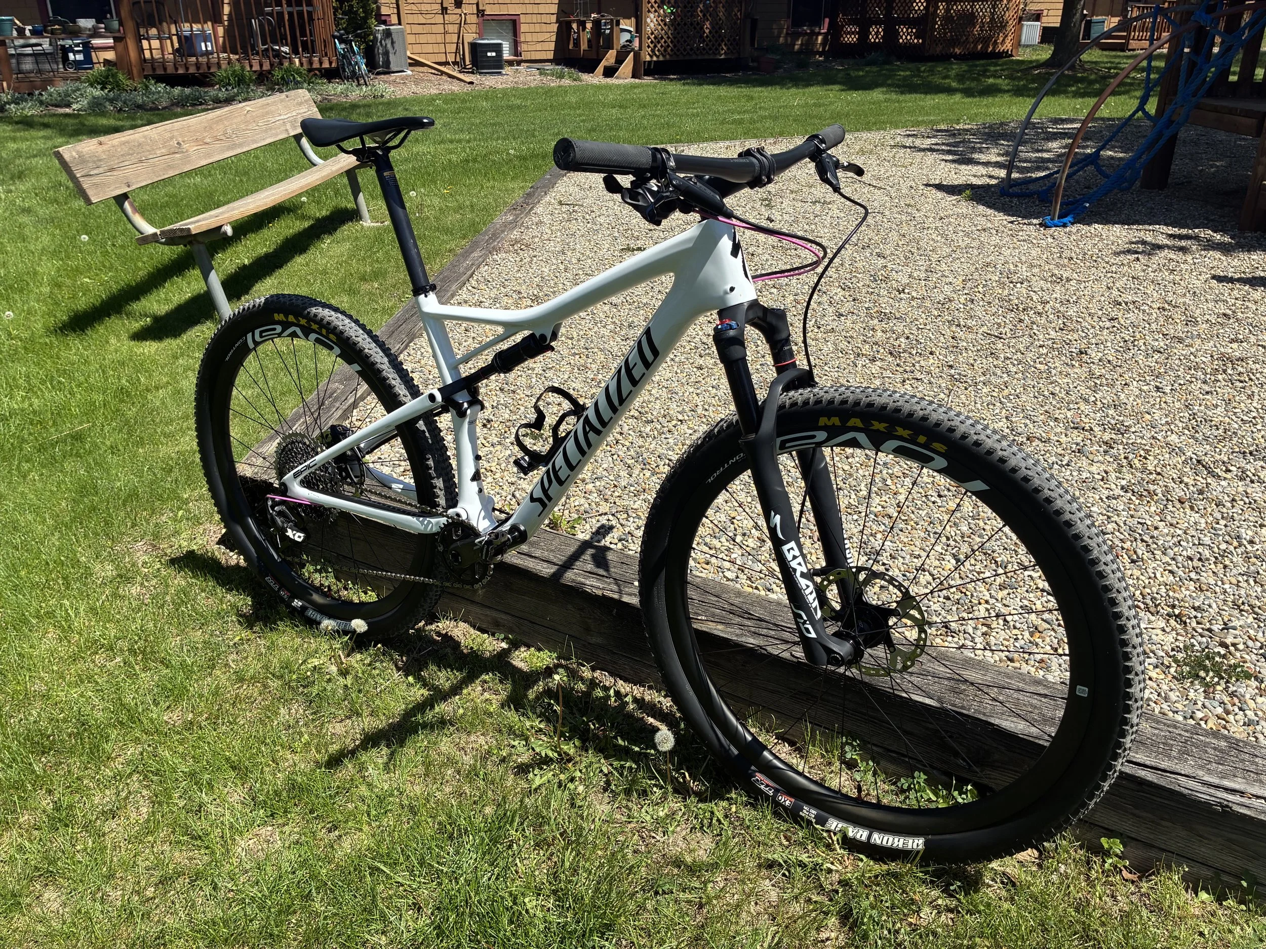 A white Specialized mountain bike with black and pink details is leaning against a wooden border on a gravel playground surface. There is a wooden bench, green grass, a playground structure with blue climbing ropes, and a wooden building with a deck 