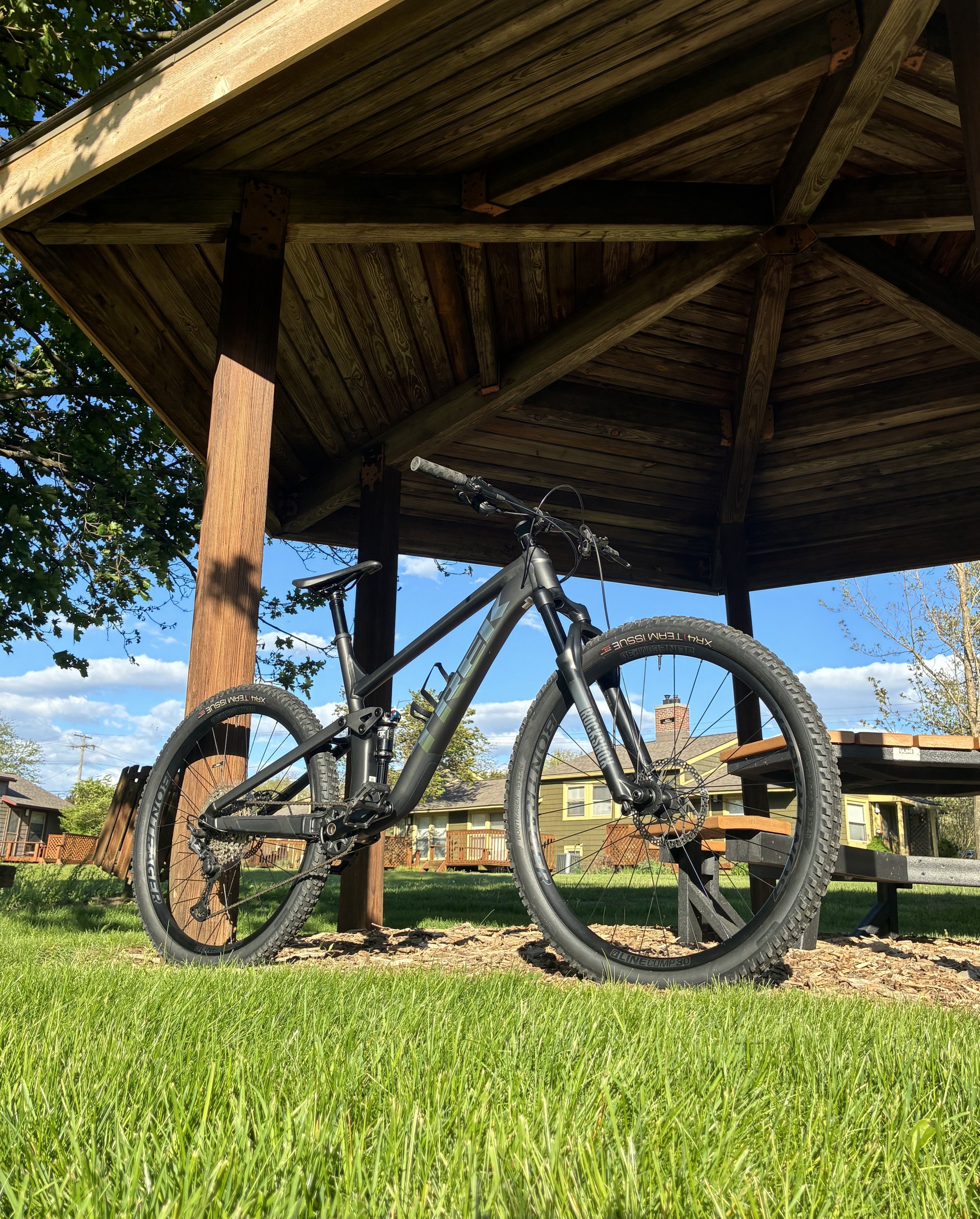 Mountain bike under a wooden gazebo in a backyard with green grass and a house in the background.