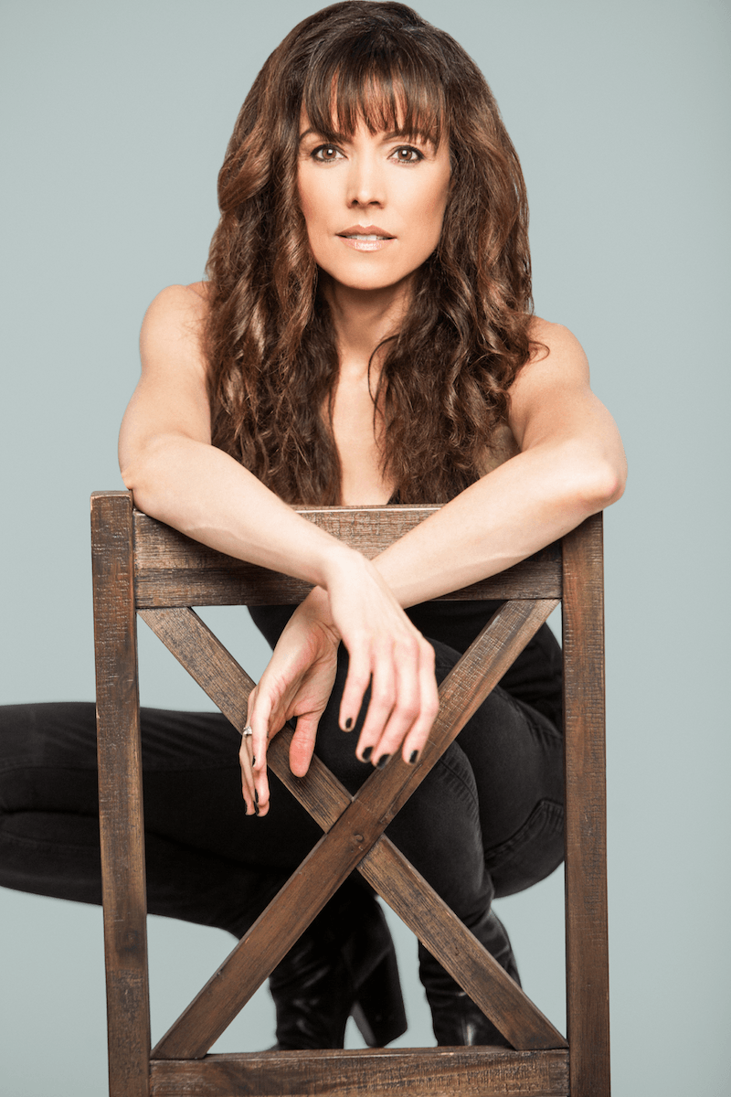 A woman with long, wavy brown hair sitting on the floor behind a wooden chair, resting her arms on the top of the chair, wearing a black sleeveless top and black pants, with a light gray background.