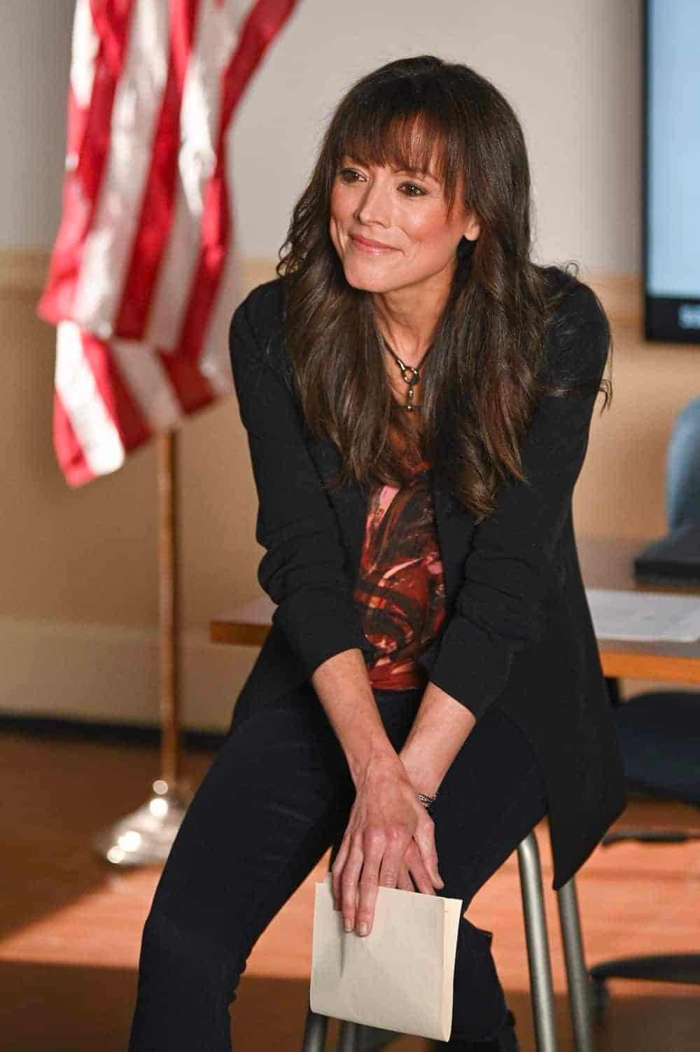 A woman in a black blazer and red shirt sitting on a stool, holding a piece of paper, with an American flag in the background.