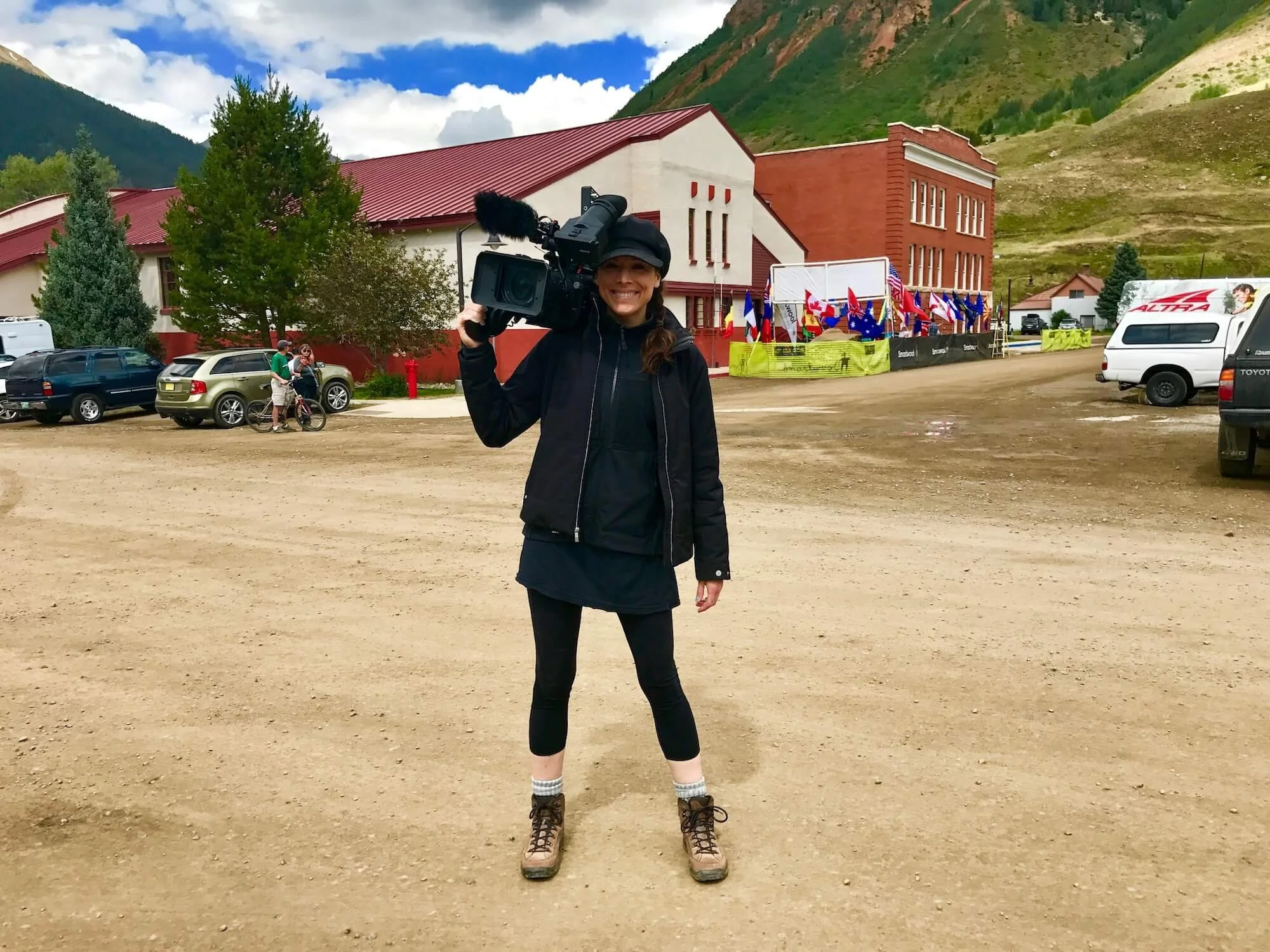 A woman smiling and holding a large video camera outdoors in a mountainous area. She is dressed in a black jacket, black cap, black leggings, and tan hiking boots. Behind her, there are parked cars, a building with a red roof, flag banners, and a backdrop of green mountains and a partly cloudy sky.