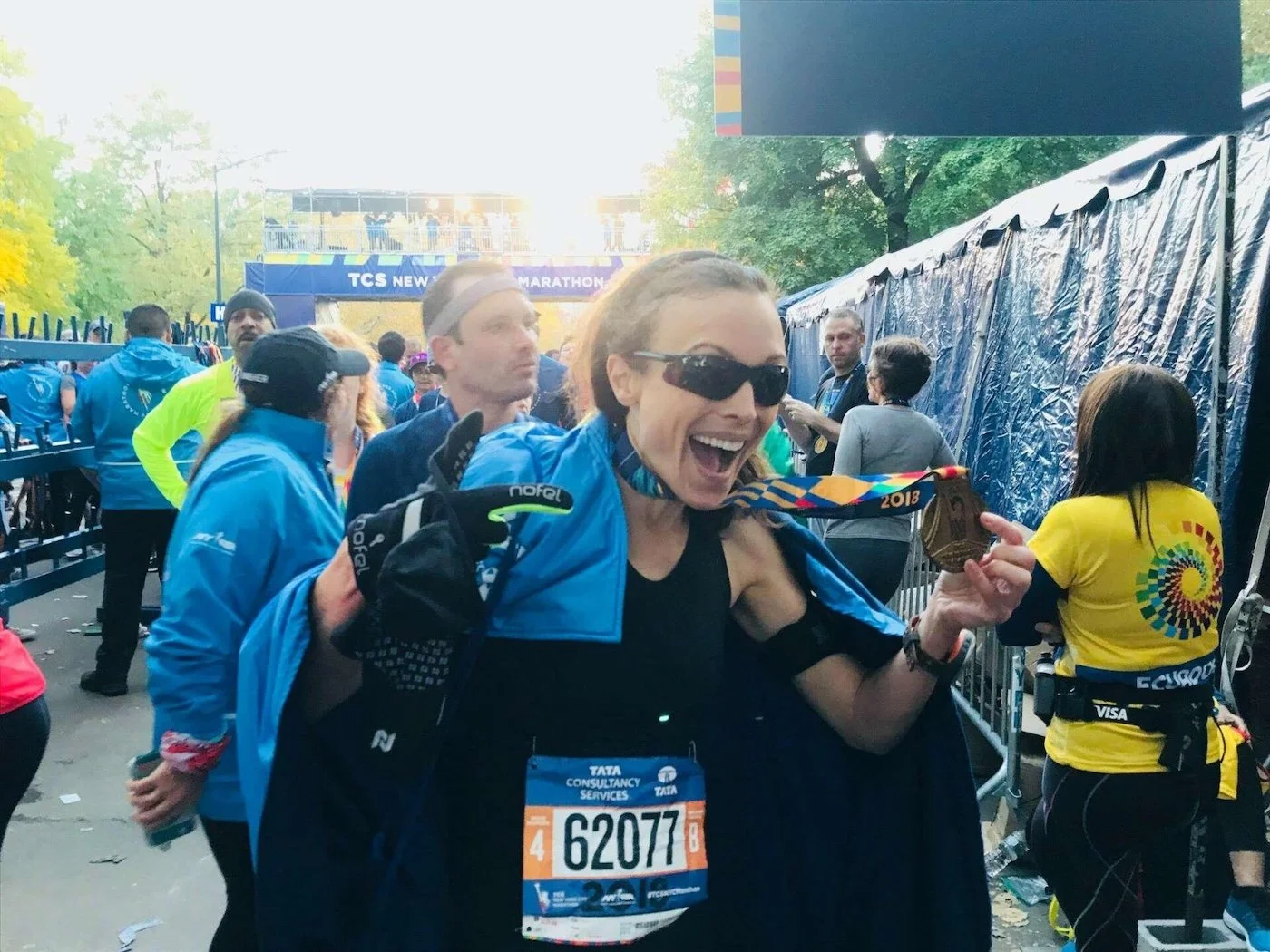 A woman wearing sunglasses and a black athletic outfit is holding a medal and smiling at a marathon event. She has a blue jacket draped over her shoulders, and a race bib numbered 62077 is pinned to her shirt. The background shows other runners, event staff, and a finish line banner that reads 'TCS New York City Marathon'.