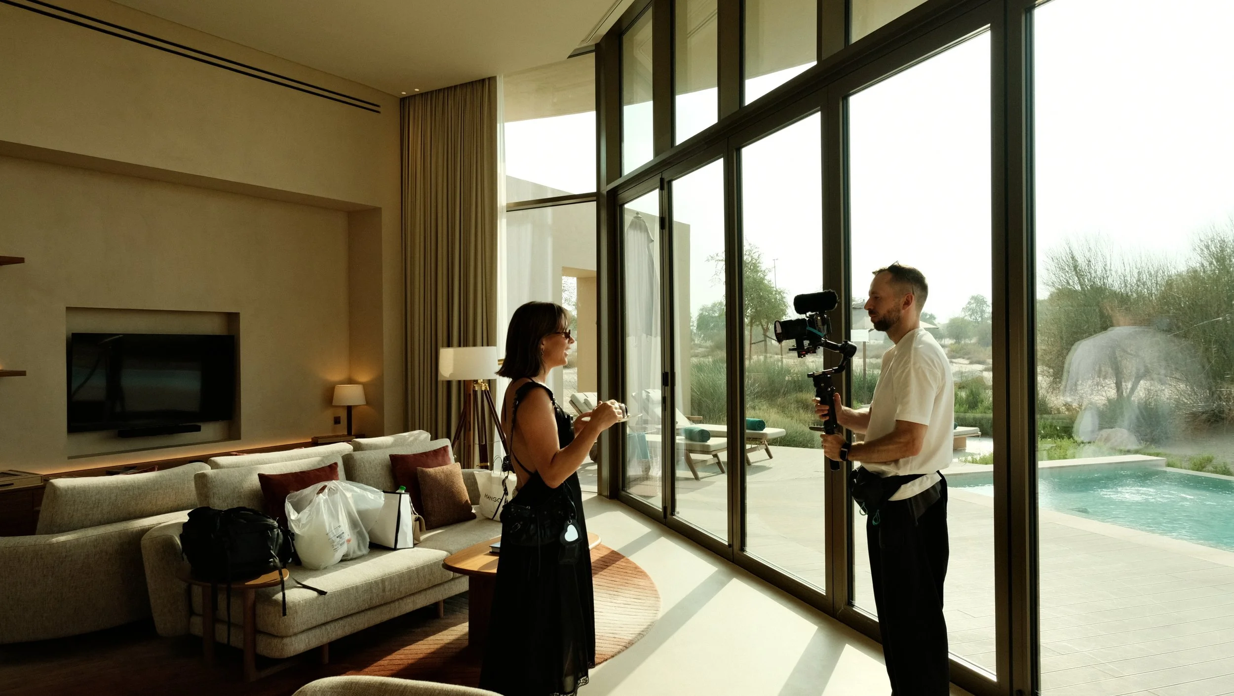 A woman holding a Small Clear Ice Cube while talking to a man with a camera inside a modern living room with large glass windows and outdoor pool
