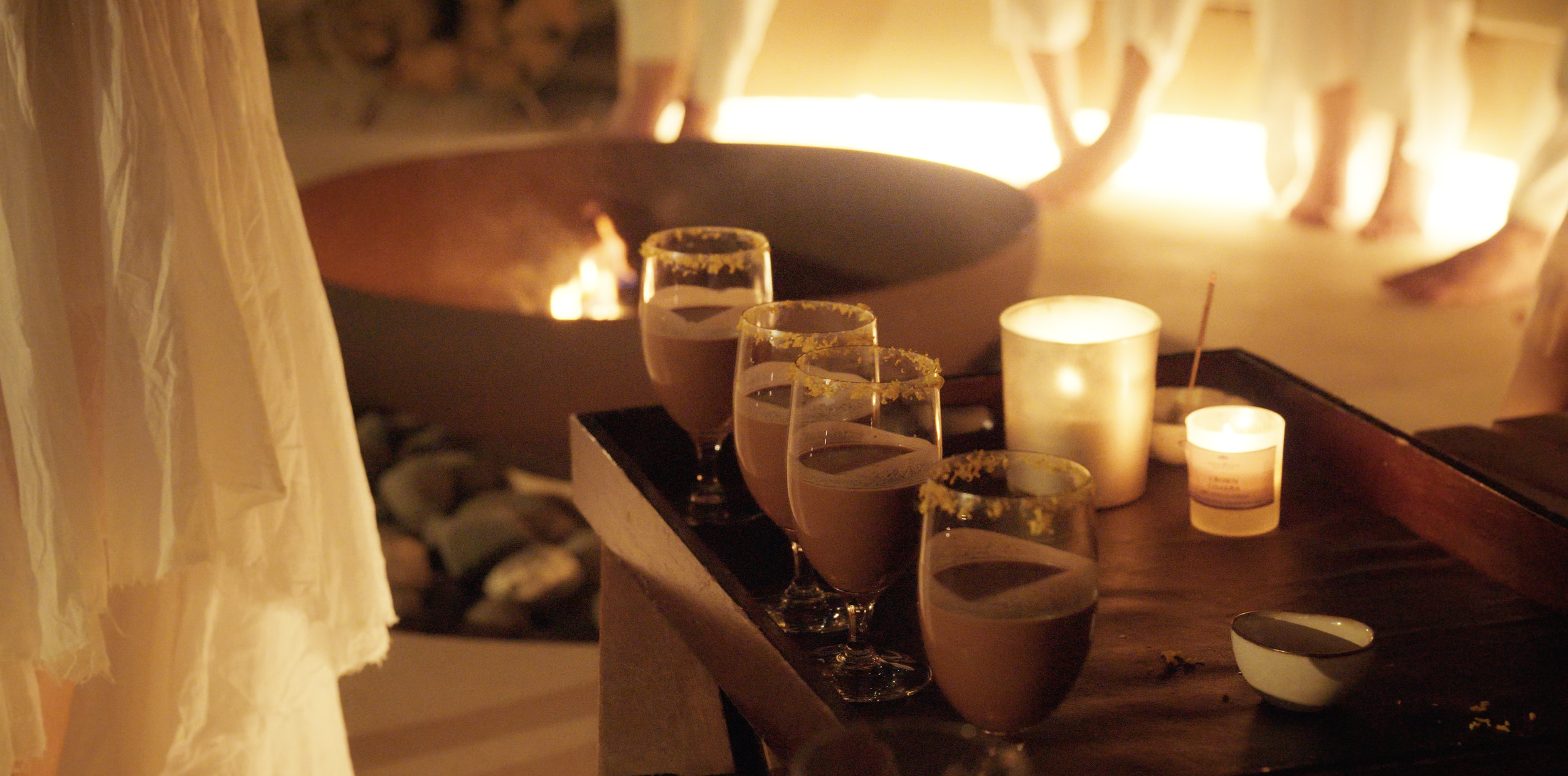 Four glasses of chocolate mousse with yellow sugar rim, candles, and a fire pit in the background, with people's feet and legs visible.