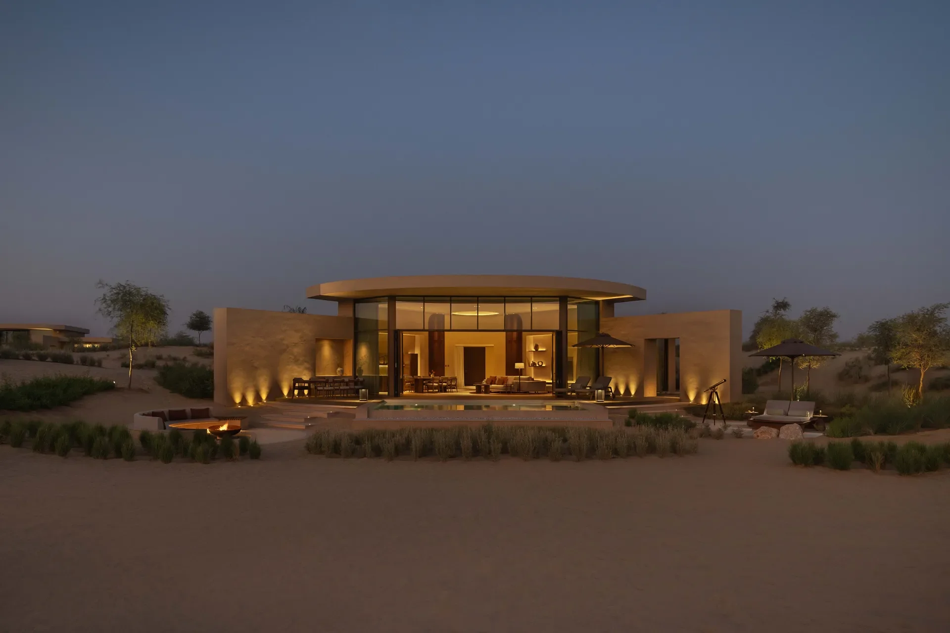 Modern house with large glass windows illuminated at dusk, surrounded by desert landscape and sparse trees.