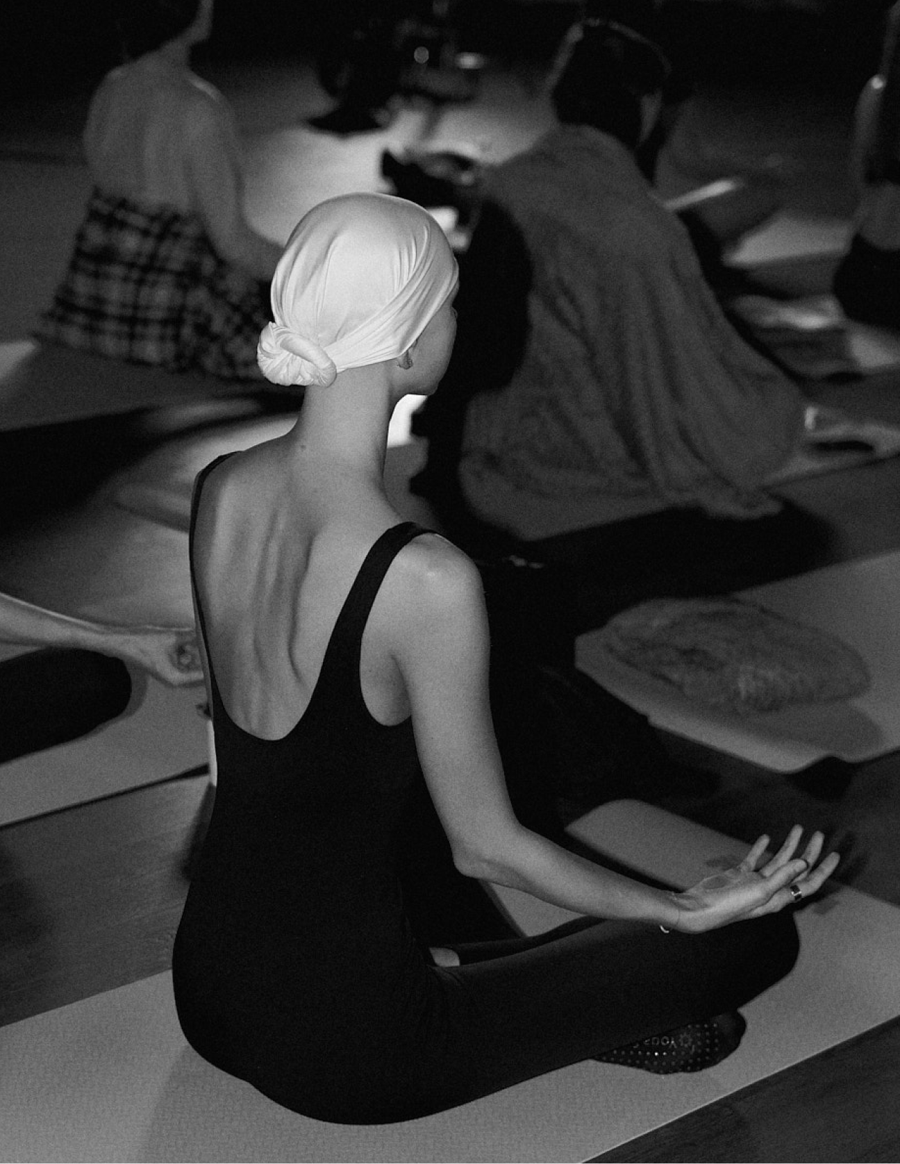A woman with a white headscarf and black tank top seated on a yoga mat, practicing meditation, in a dark room with other people sitting on mats in the background.