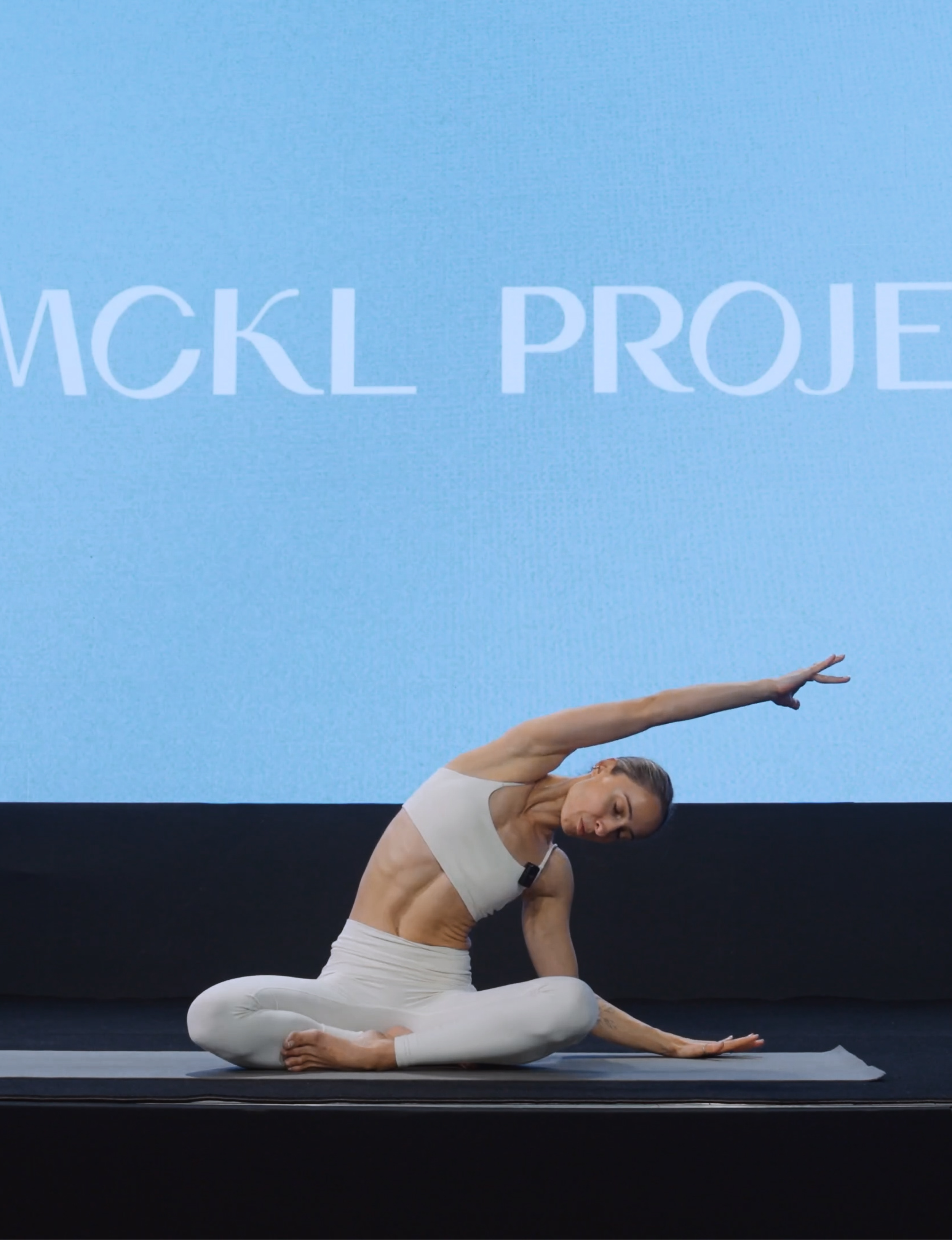 A woman in white athletic clothing performing a seated side stretch yoga pose on a mat on stage with a large screen in the background.