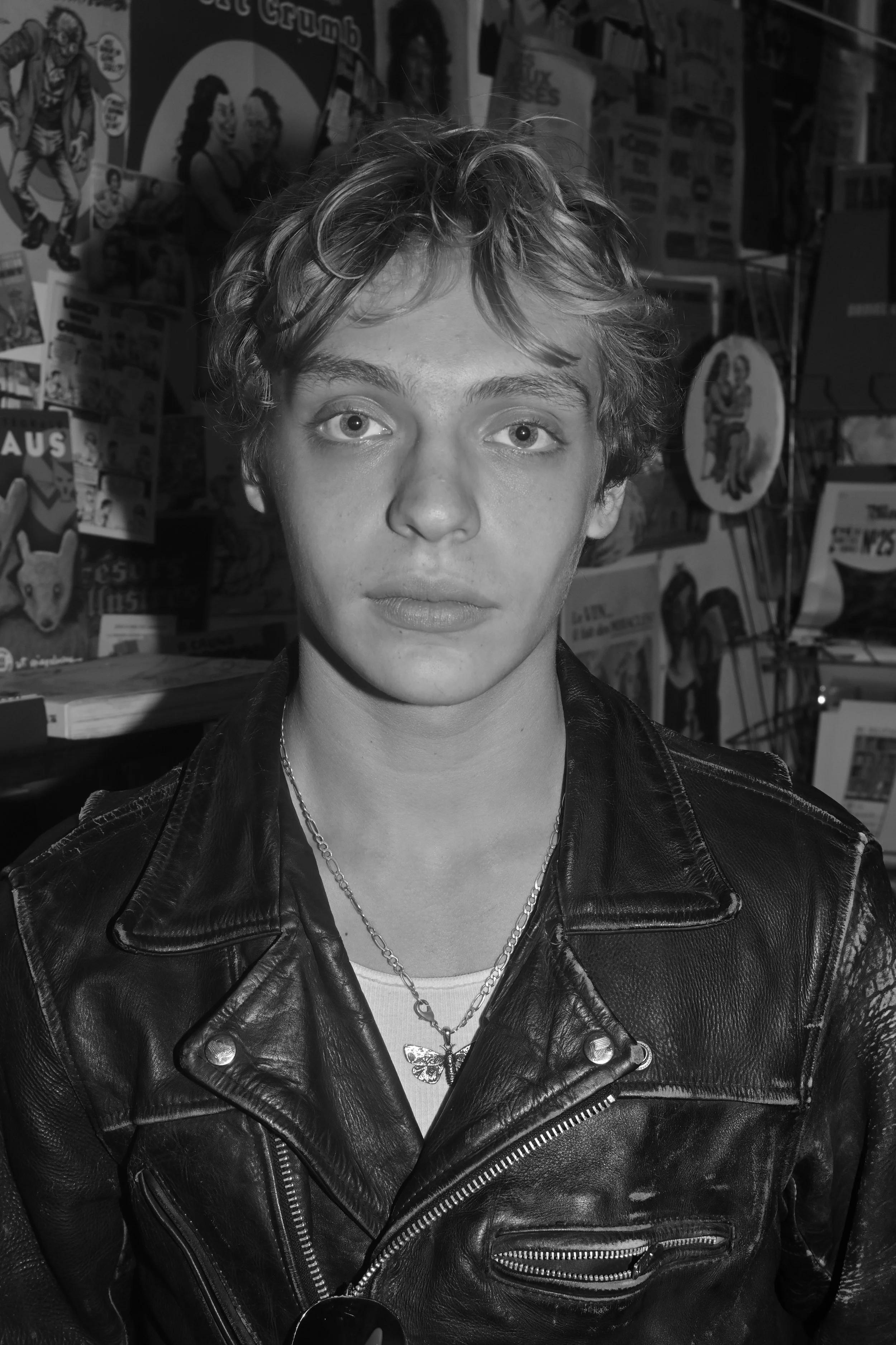 Black and white photo of a young person with curly hair wearing a leather jacket and silver jewelry, standing in a room with comic book posters on the wall.