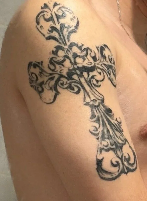 A black tribal-style tattoo of a sword with ornate, swirling patterns on a person's upper arm and shoulder.