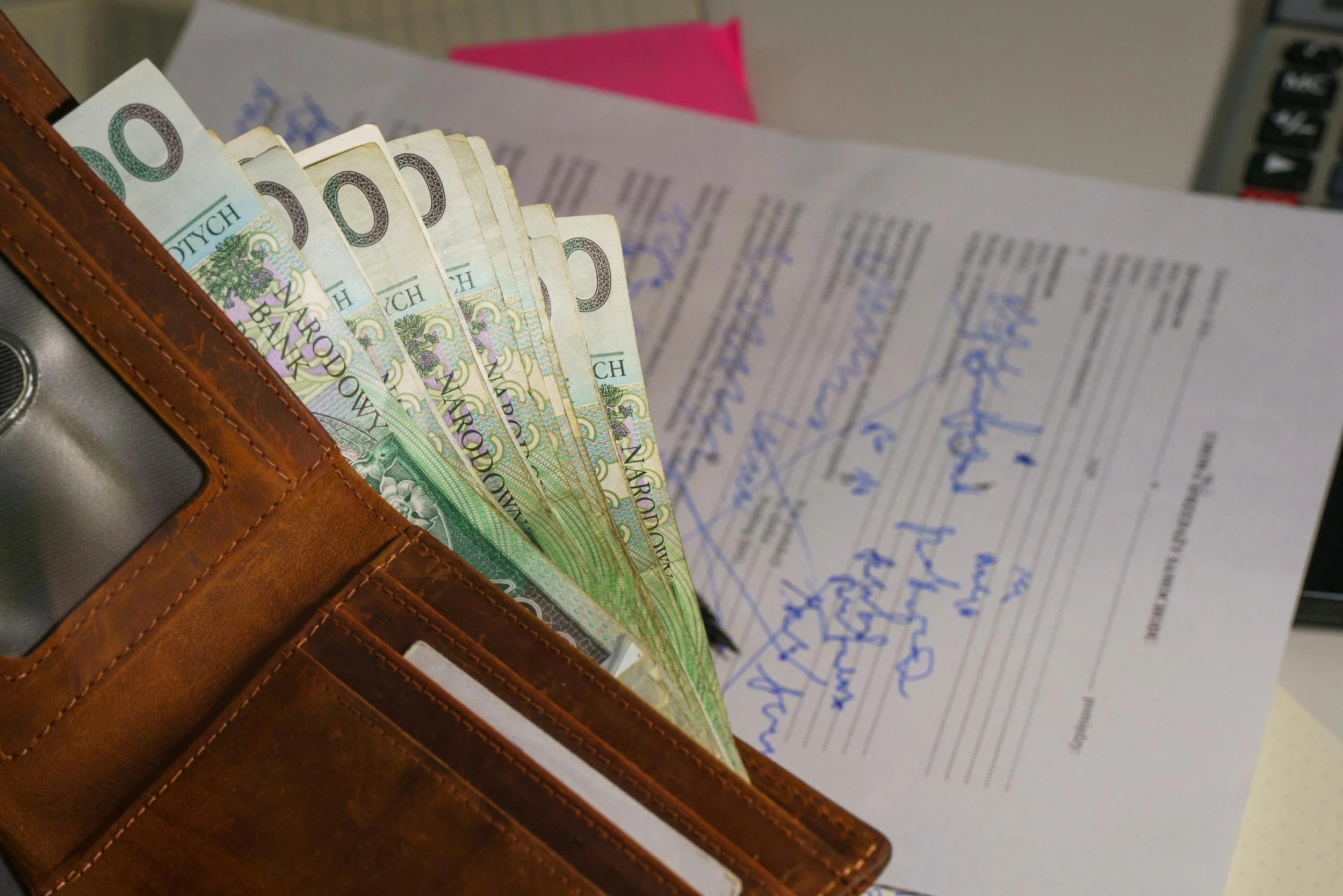 A leather wallet holding several Polish 20 zł banknotes, placed on top of handwritten notes with blue ink.