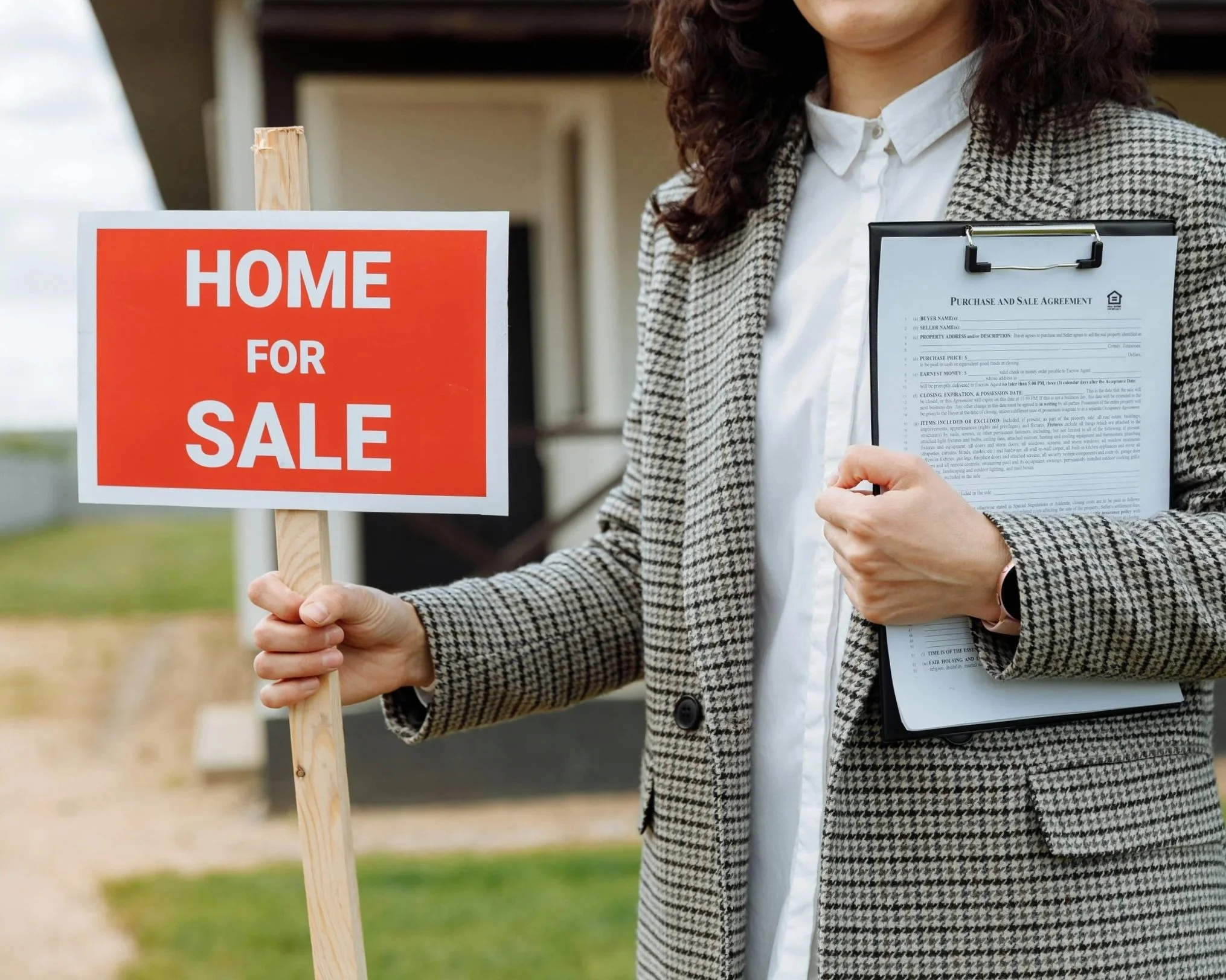 A woman holding a wooden sign that says 'Home for Sale' and a clipboard with a purchase agreement, standing outdoors.