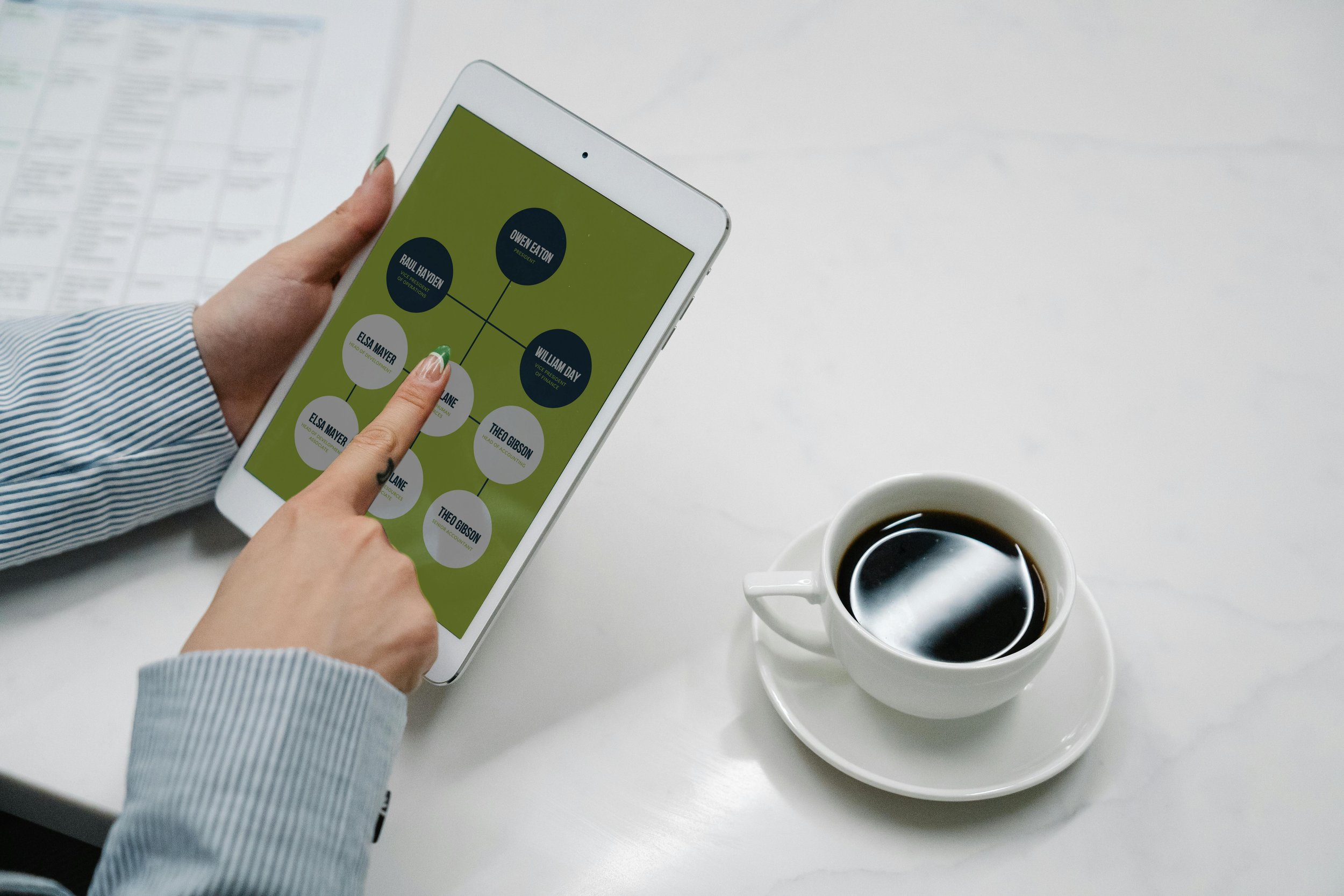 Person holding a tablet displaying a chart with names and roles, next to a cup of coffee on a white table.