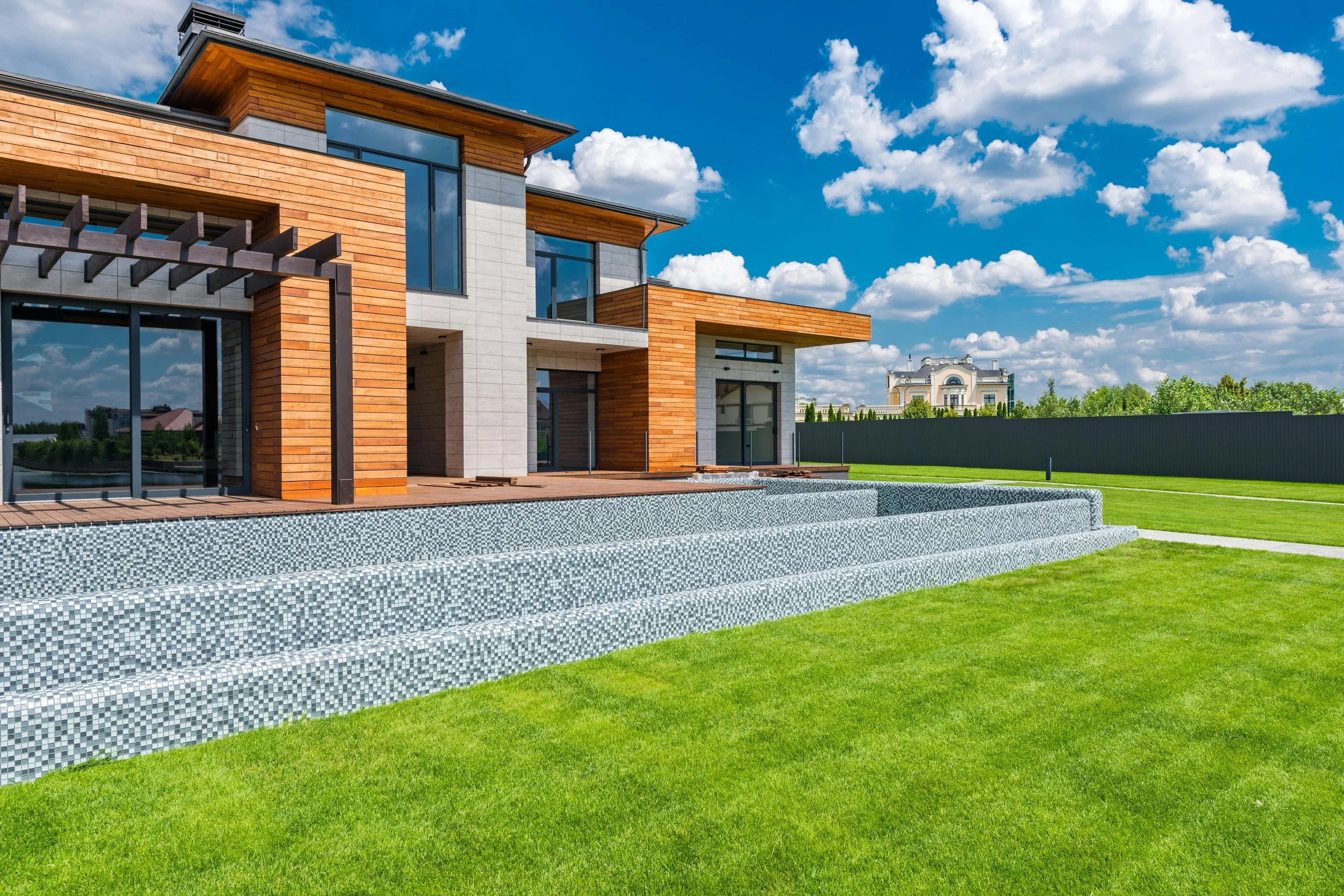 Modern house with large windows, wooden siding, and gray concrete accents, surrounded by a green lawn and set against a blue sky with clouds.