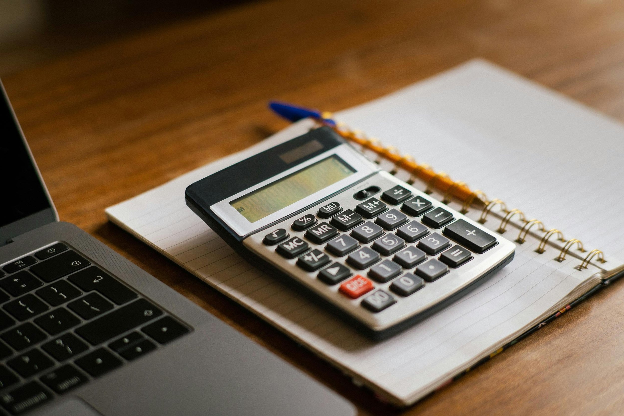 Calculator, notebook, blue pen, and a laptop on a wooden desk.