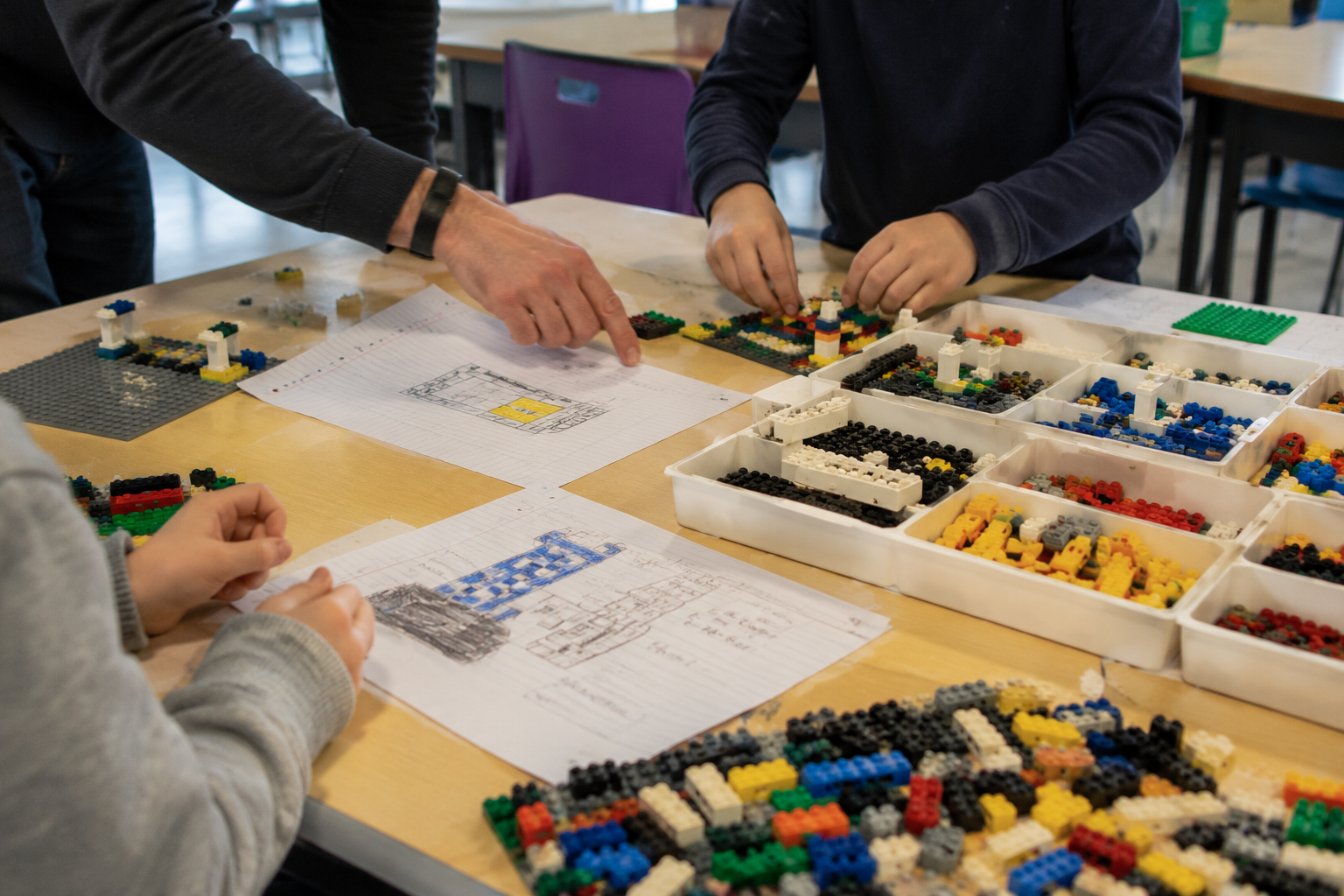 People working on a LEGO building project at a table, with LEGO pieces organized in trays, sketches, and instructions.